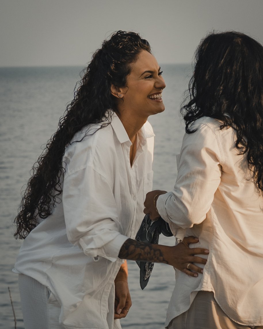 Two women face each other, one has her hand on the others hip. Her face is shown and she is smiling. 