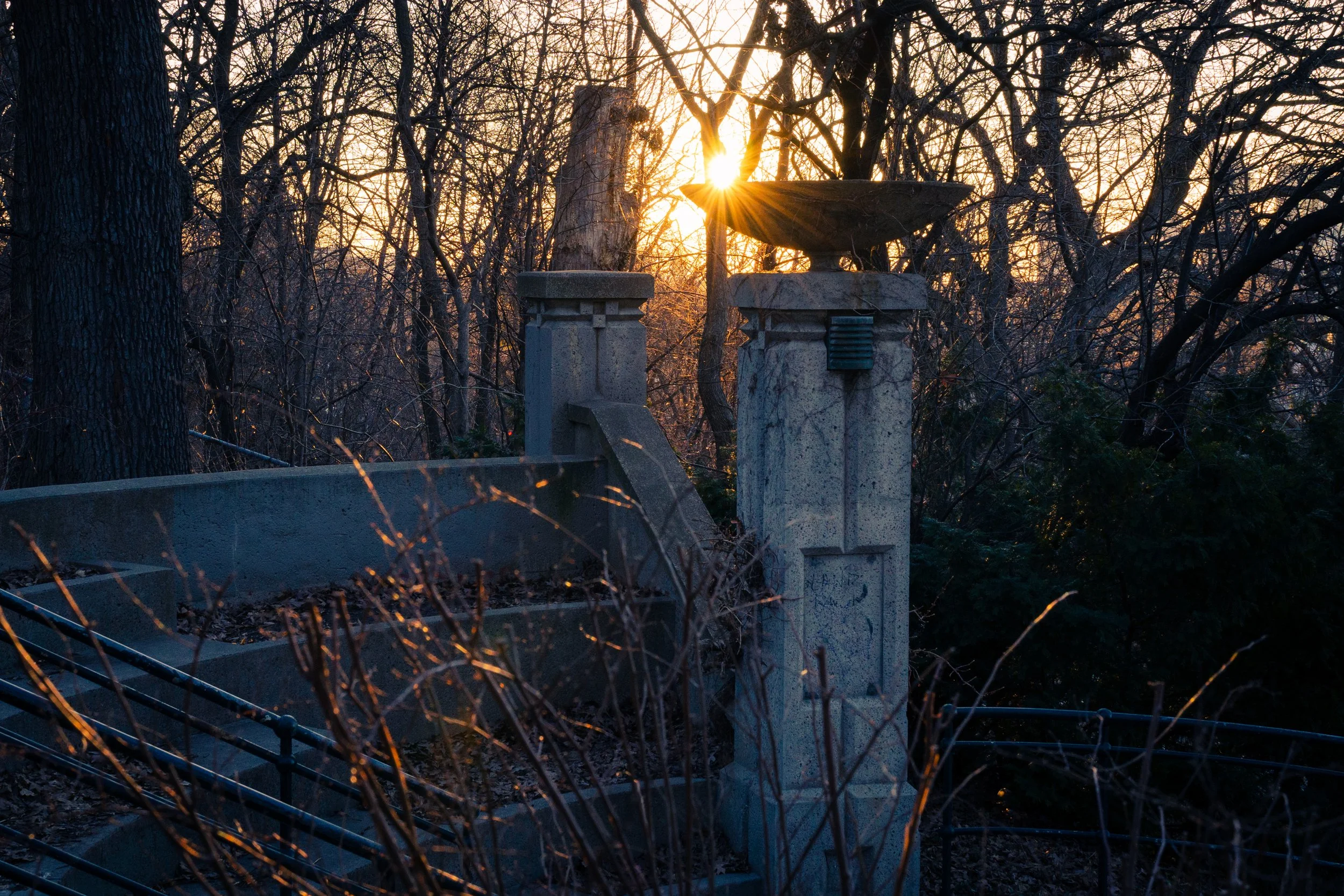 An outdoor scene with a stone column and stairs silhouetted against a setting sun surrounded by leafless trees.