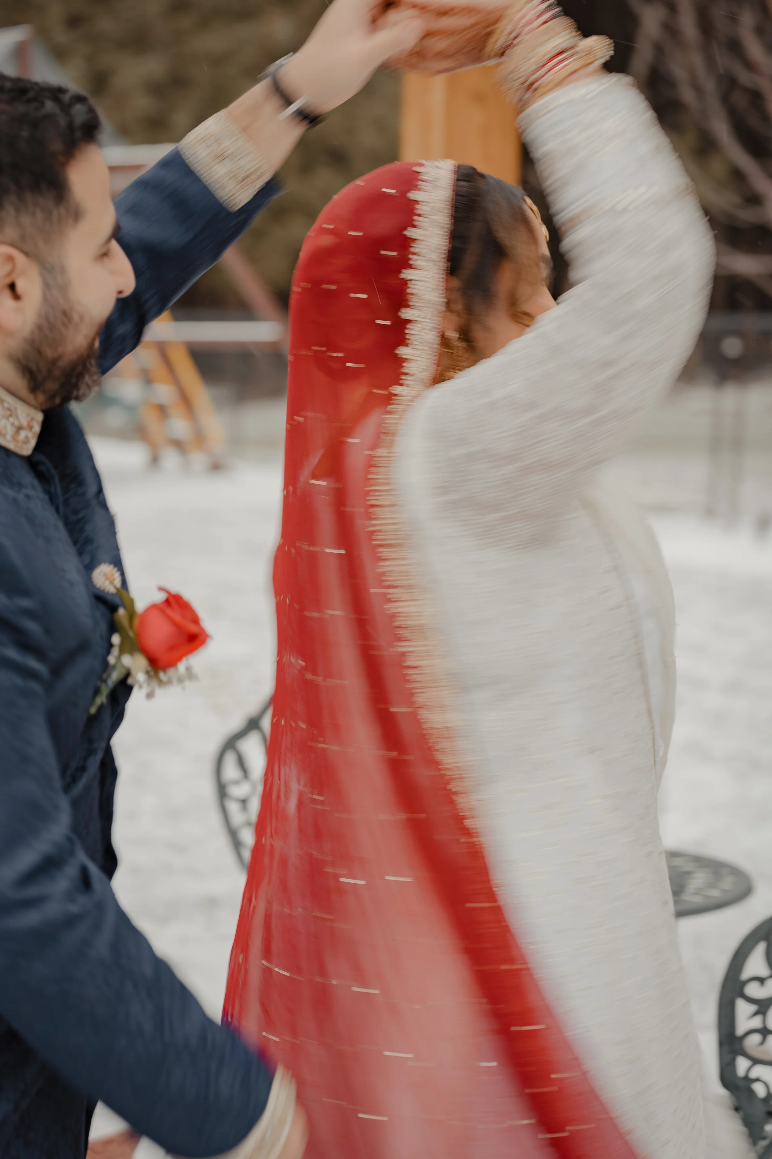 A bride being spun by her groom in a backyard with snow on the ground. She is wearing a deep red dupatta and white dress. He is wearing a dark blue qameez. 