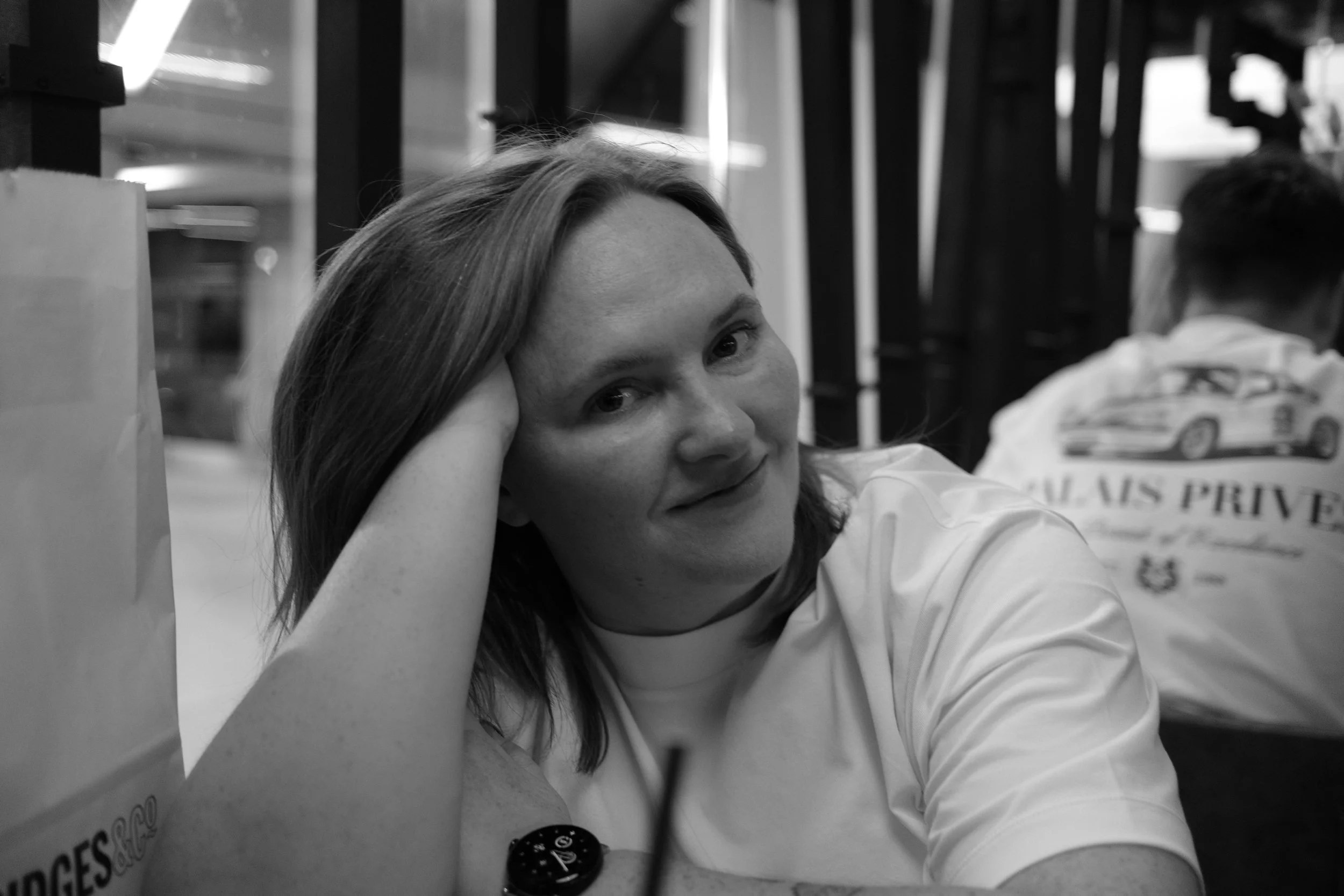 A woman with shoulder-length hair resting her head on her hand, sitting at a table in a restaurant or cafe, with a man in the background wearing a plain white t-shirt. The image is in black and white.