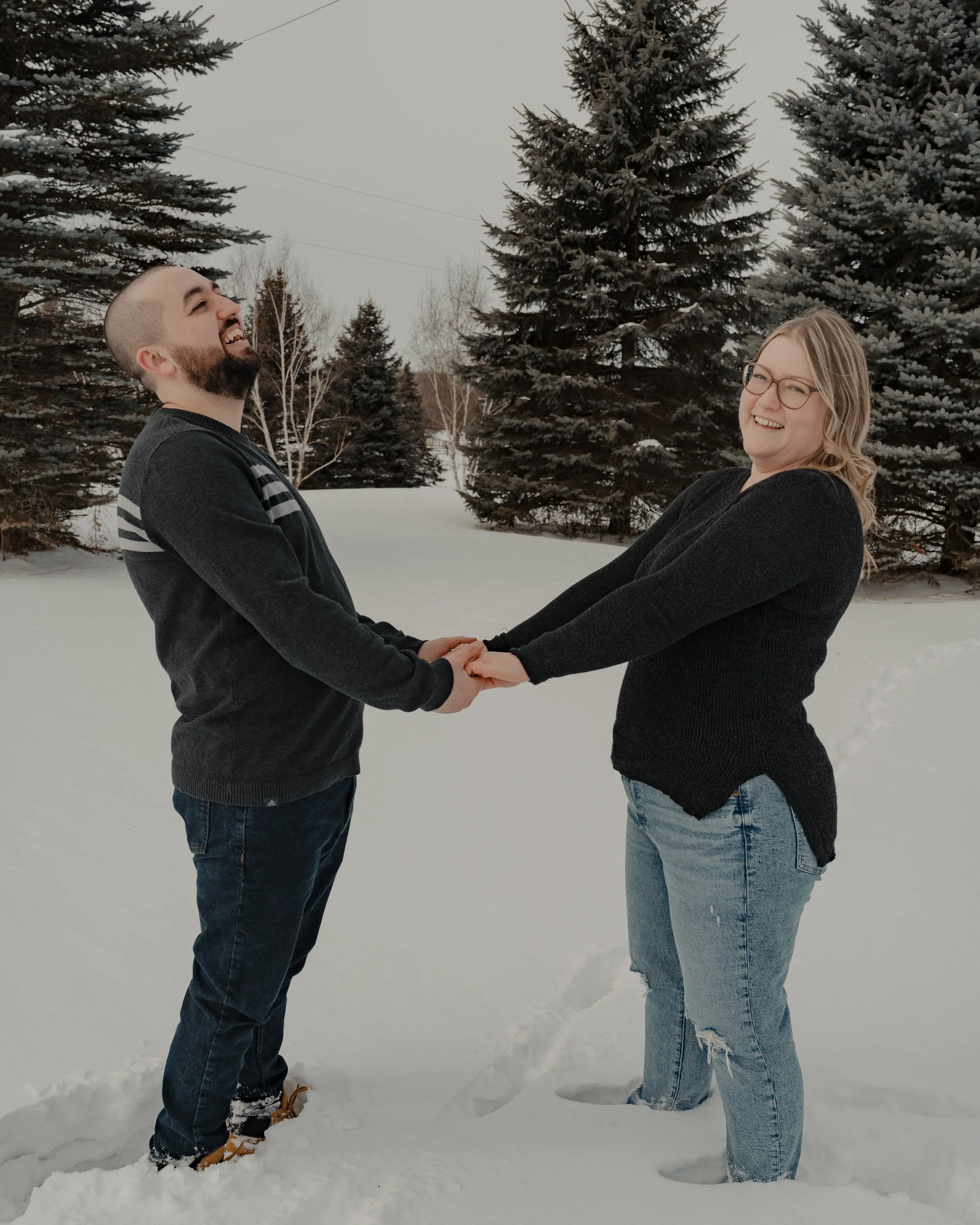 A man and a woman face each other standing in snow up to their ankles. They are holding hands and laughing. They are both wearing jeans and dark tops. 