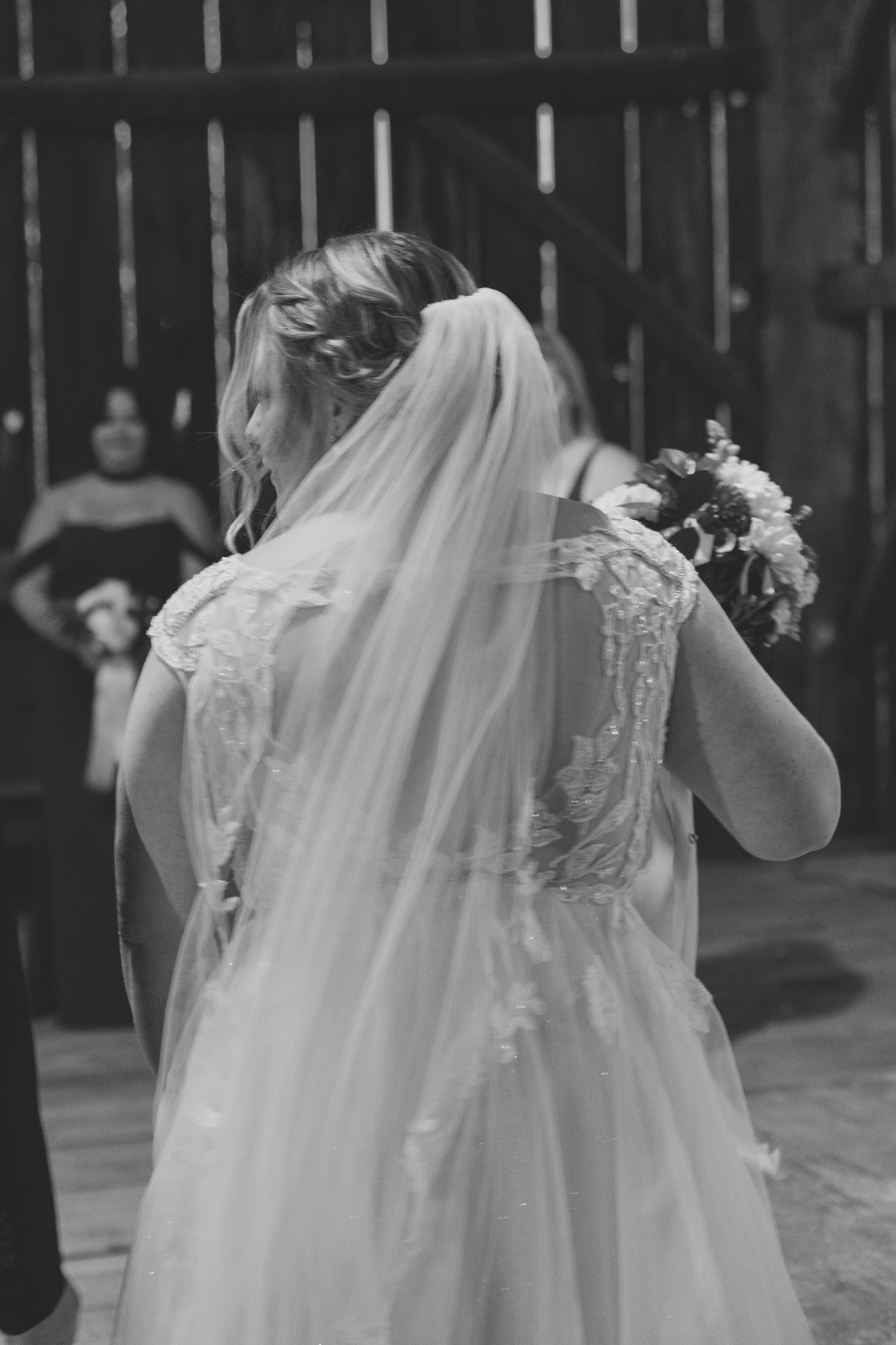 A woman is seen from behind, she is looking to the left with people behind her in the distance. She is wearing a wedding dress and veil, and holding a flower bouquet. The picture is in black and white.