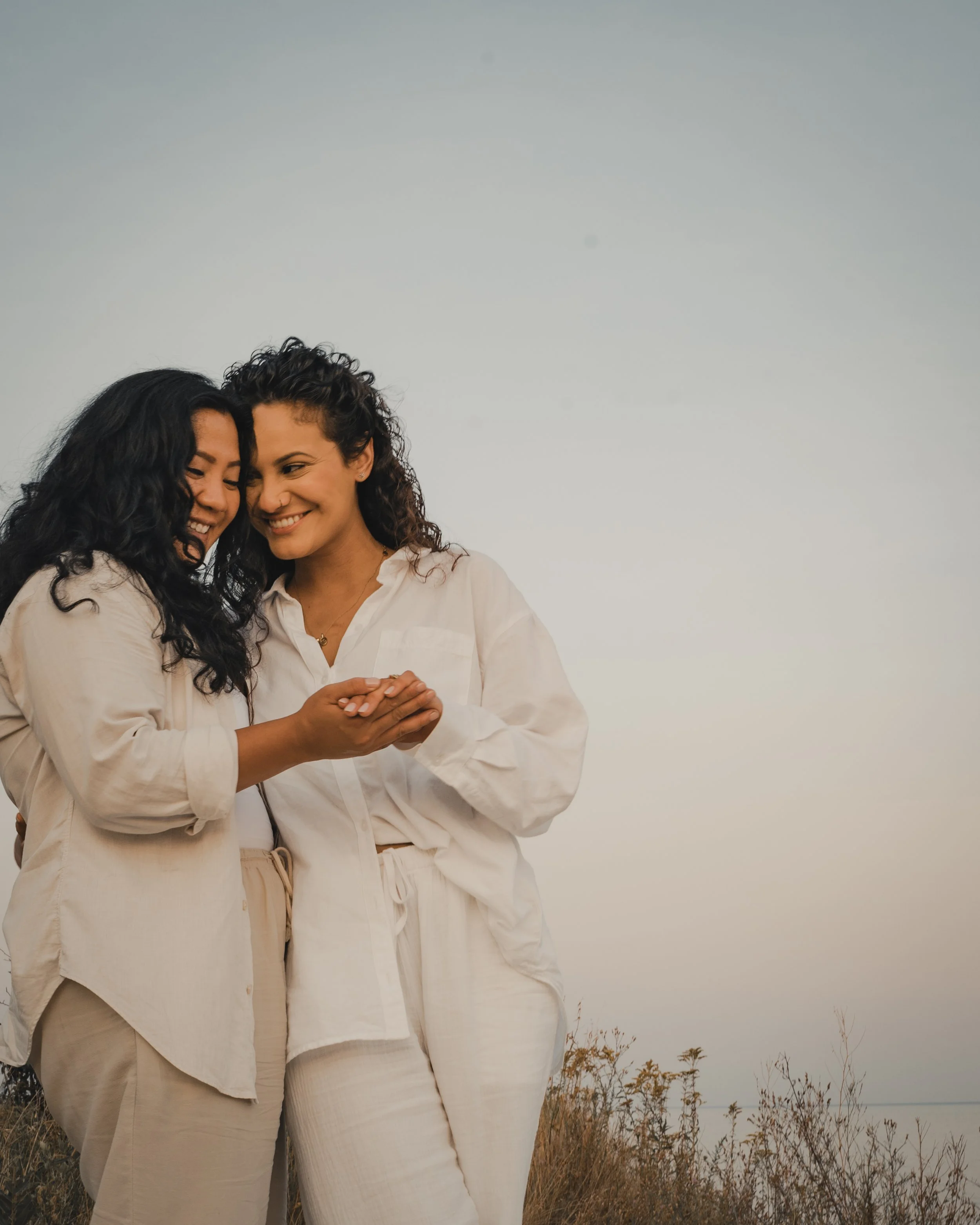 Two women stand side by side, side hugging and holding hands. They are both wearing white shirts and light pants. The sky is in the background. 