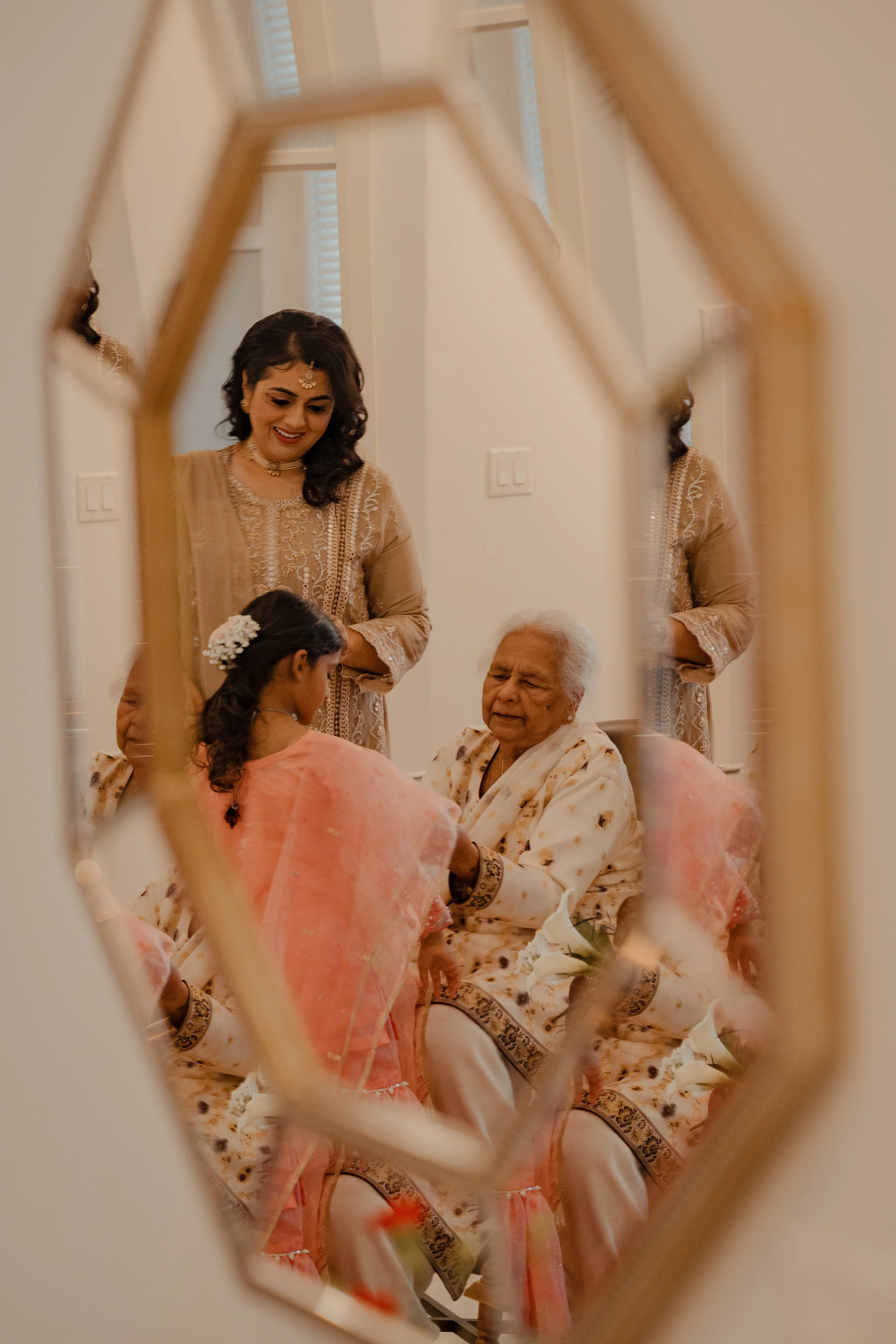 A child has her back to the viewer. She is wearing a peach coloured dupatta and showing it to older woman who is sitting and wearing a cream coloured dupatta. They are both being watched over by a woman wearing a gold coloured kurta. 
