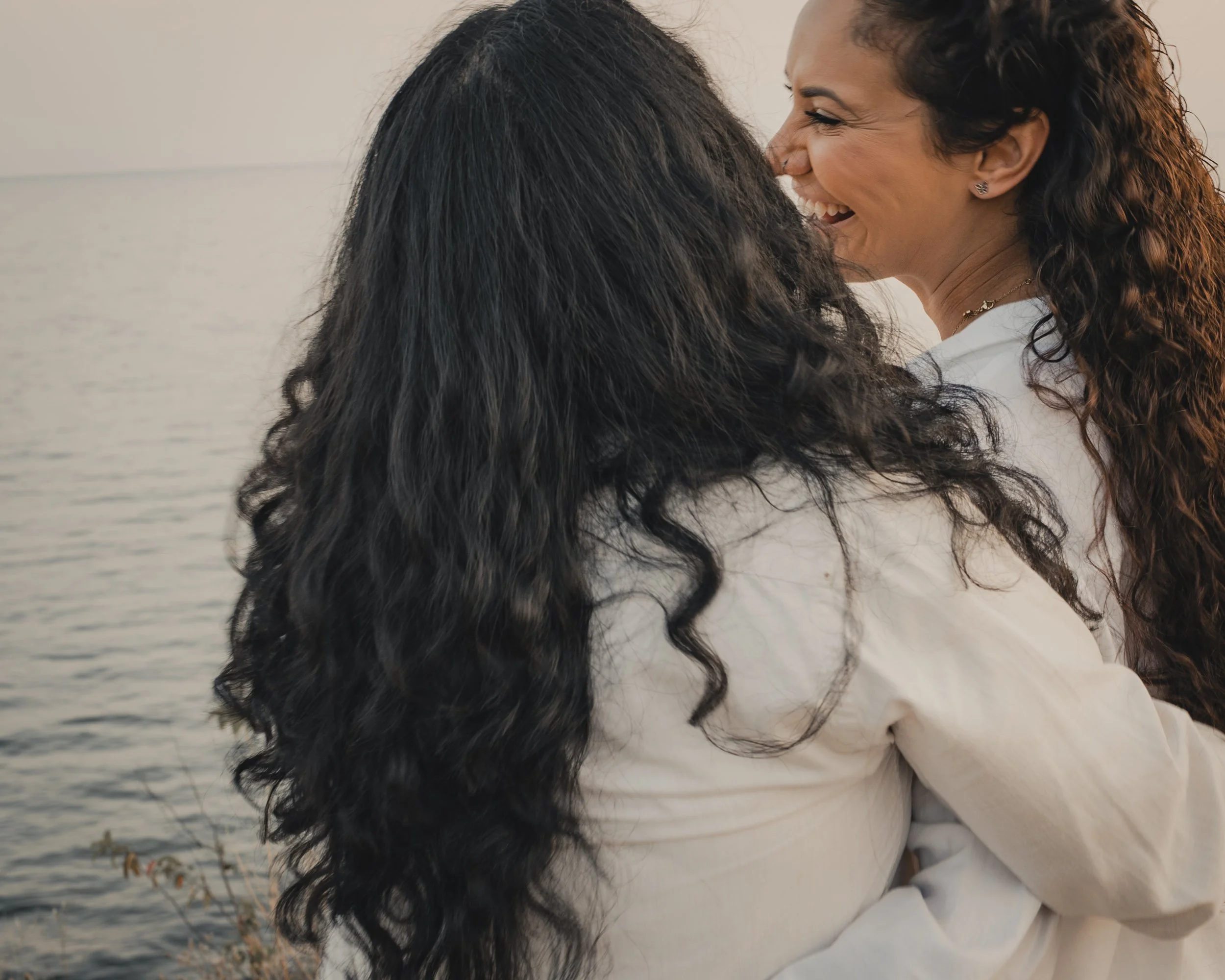 Two women face away, towards water. They are looking at each other. They are both wearing white. 