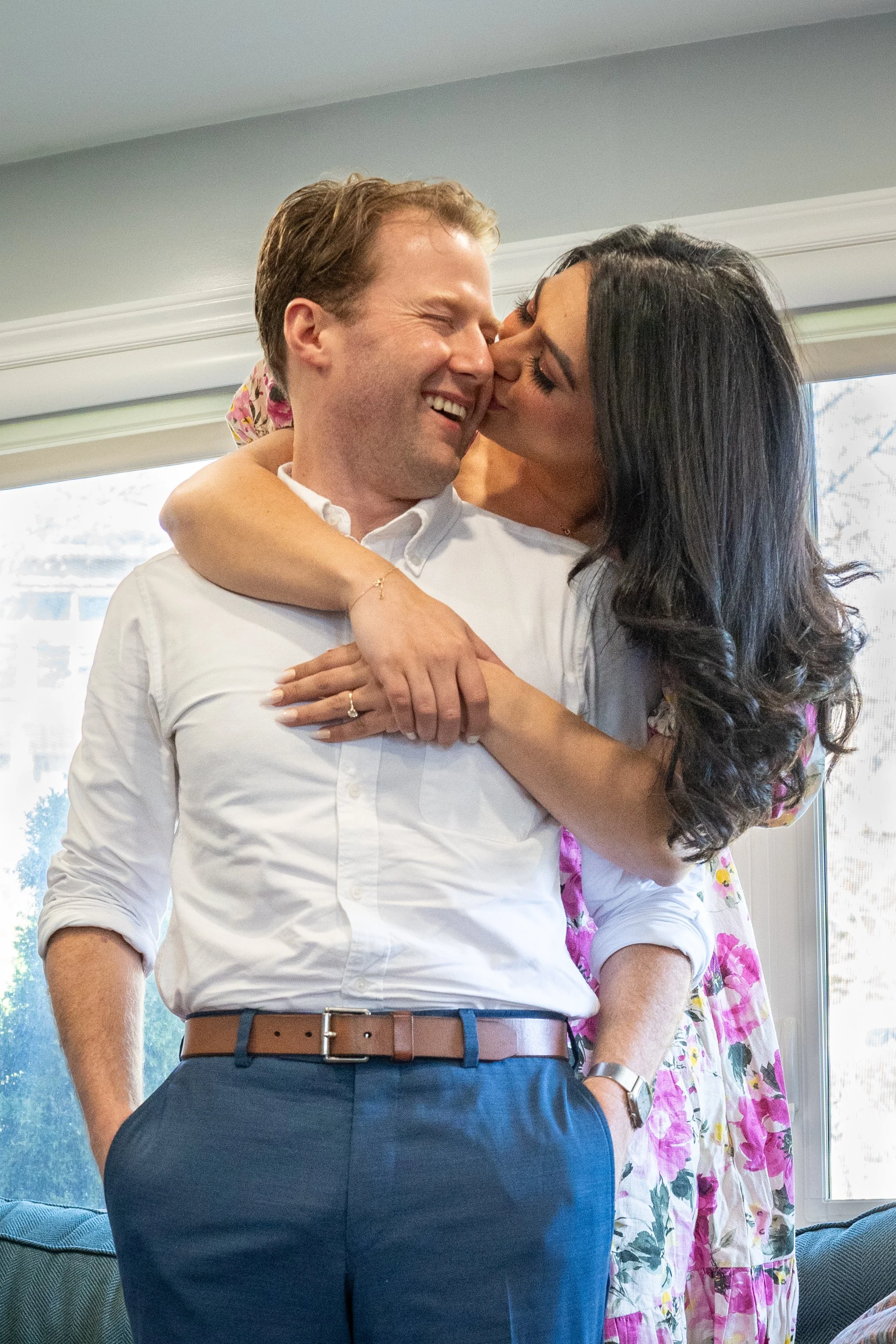 A woman stands behind a man, her arms are hugging him, she is kissing him on his right cheek. He is facing her and smiling. He is wearing a white shirt. She is wearing a floral dress. 