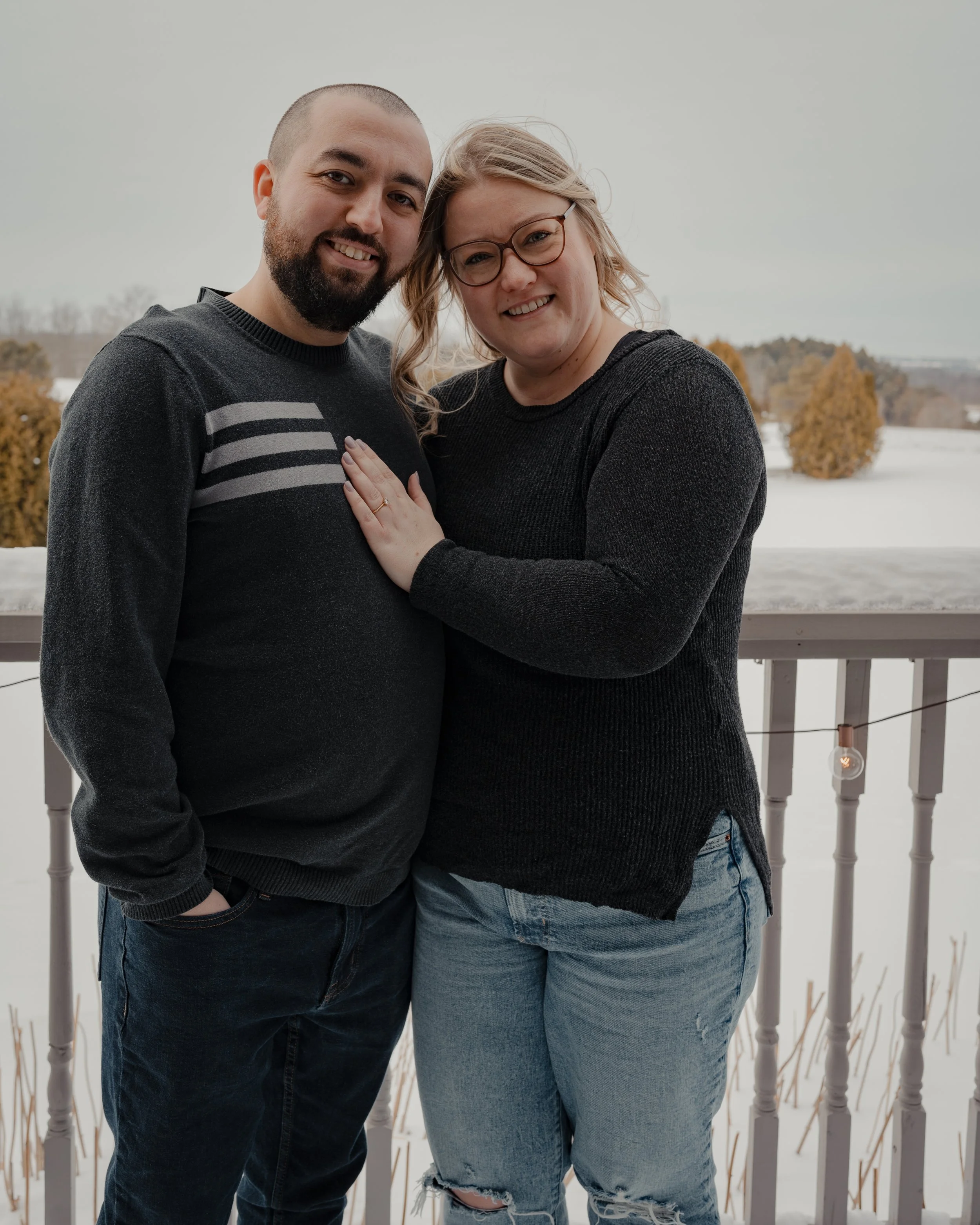 A man and woman stand side by side, side hugging looking at the camera. The woman has her hand on his chest, showing an engagement ring. There is a balcony railing and snowy field behind them. 