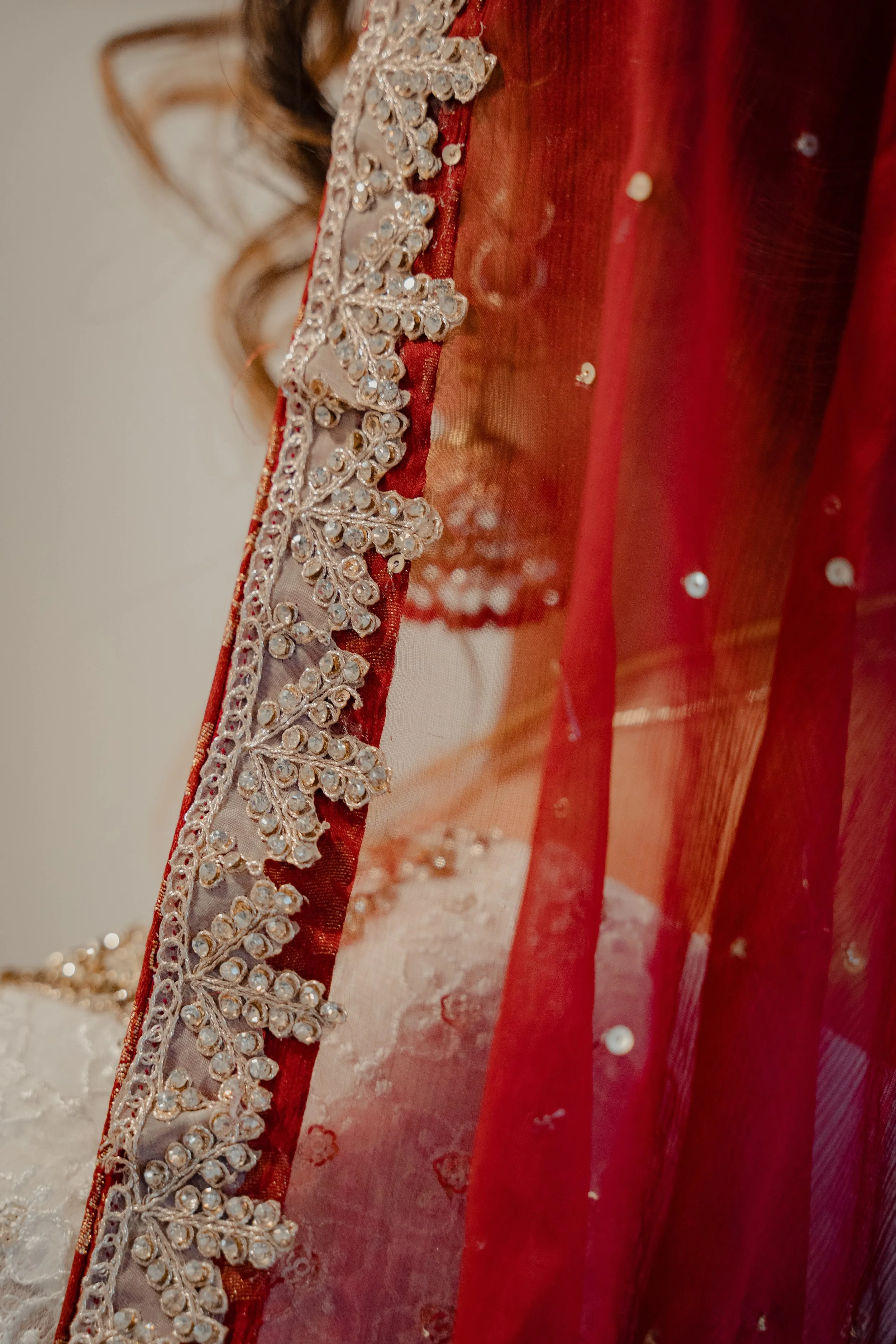 a close up of the bead work on a dark red dupatta being worn by a woman wearing a white dress and jhumka earrings which can be seen through the dupatta. 