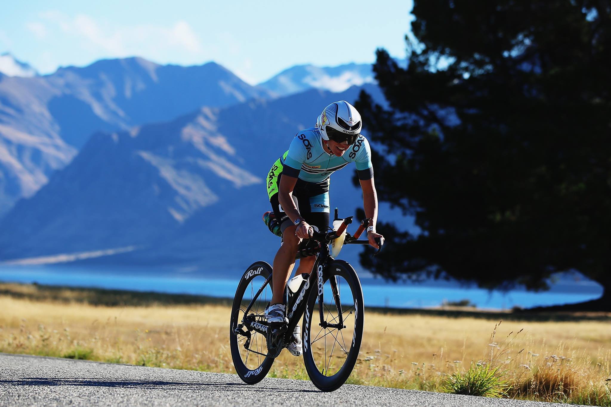 A cyclist riding a black road bike on a scenic outdoor road with mountains, a lake, and trees in the background.