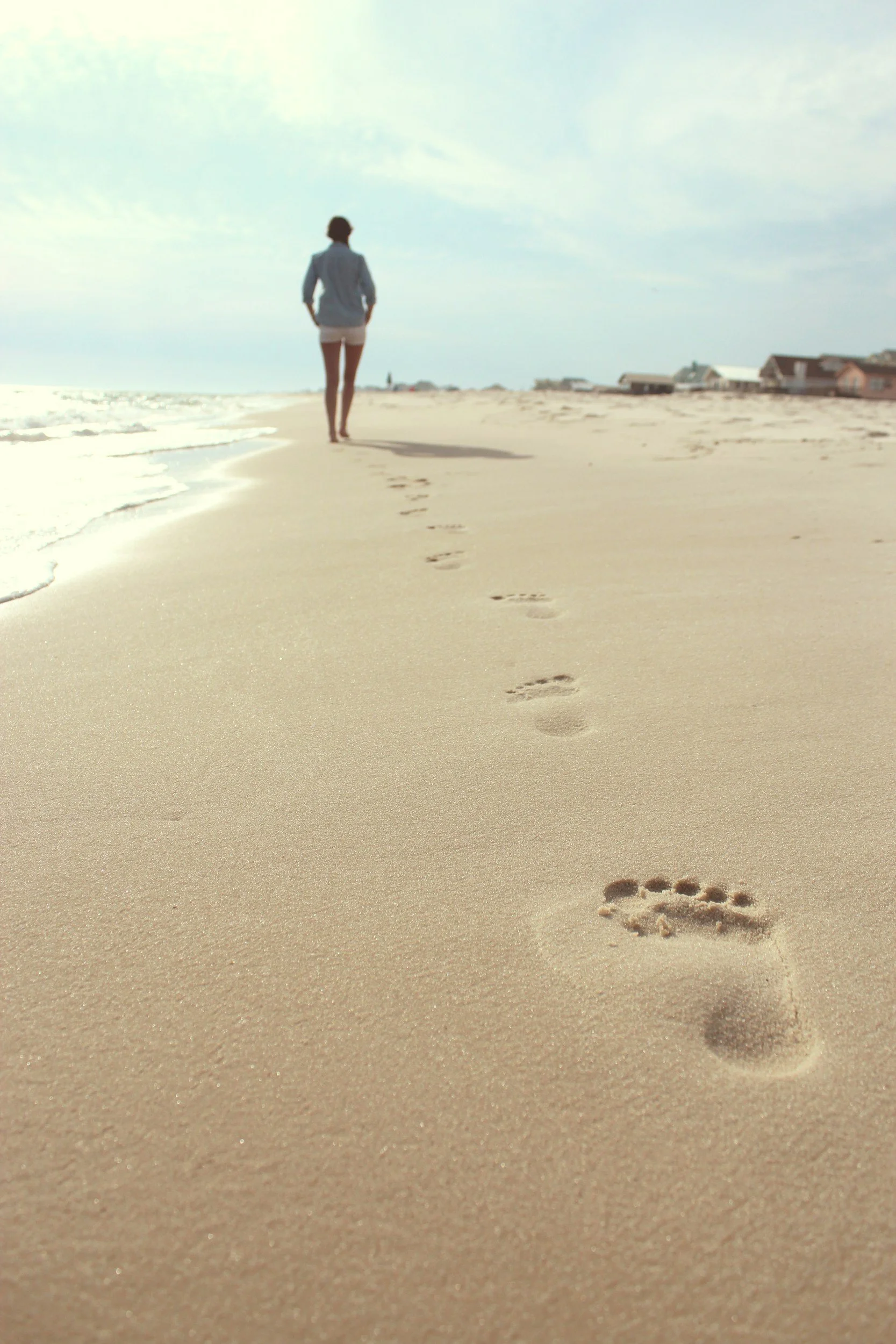 Woman walking away from the camera down the beach along the shoreline. You can see her footprints in the sand and the waves lapping the shore