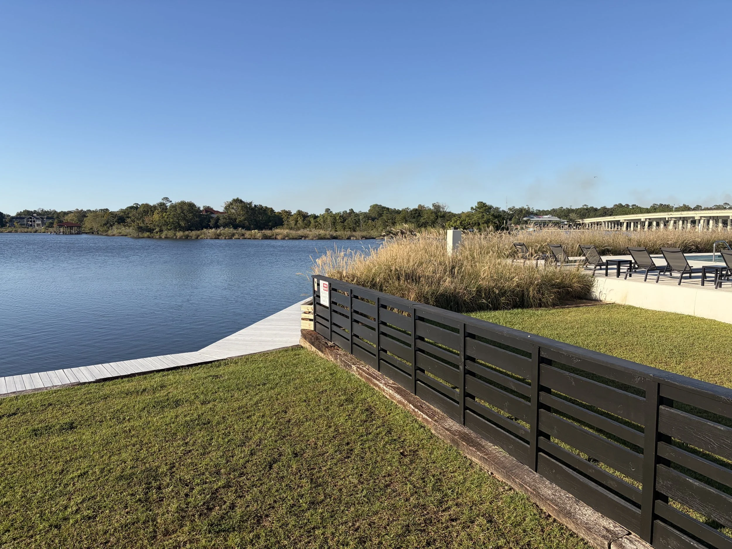 A bay view outdoor area with a wooden dock extending into the water, surrounded by grass, ornamental grasses, and seating with lounge chairs, under a clear blue sky.