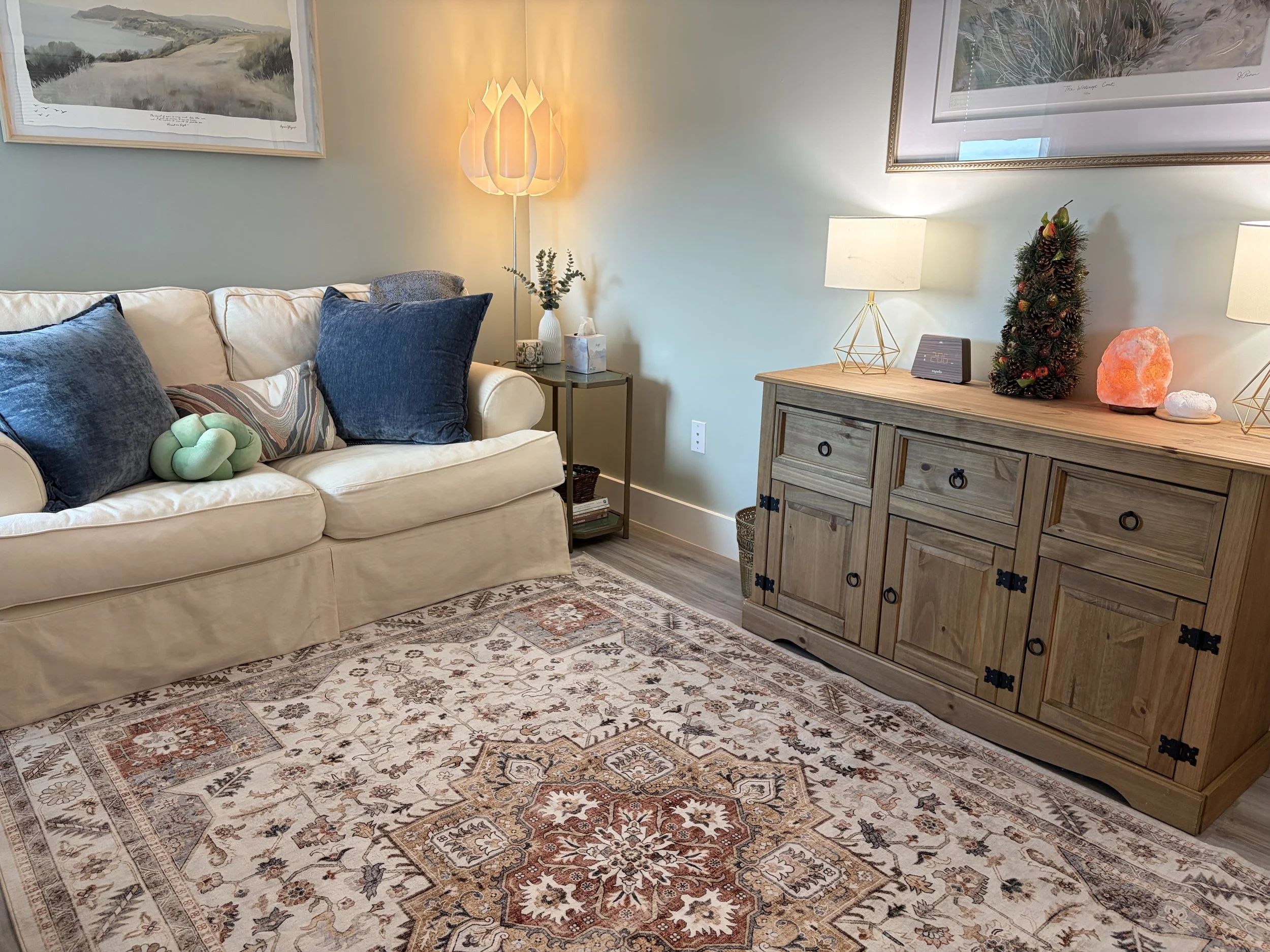 Cozy therapy room with a cream colored sofa, dark blue pillows, a patterned rug, a wooden sideboard decorated with a small Christmas tree, salt lamp, and lamps, with framed landscape artwork on the wall.