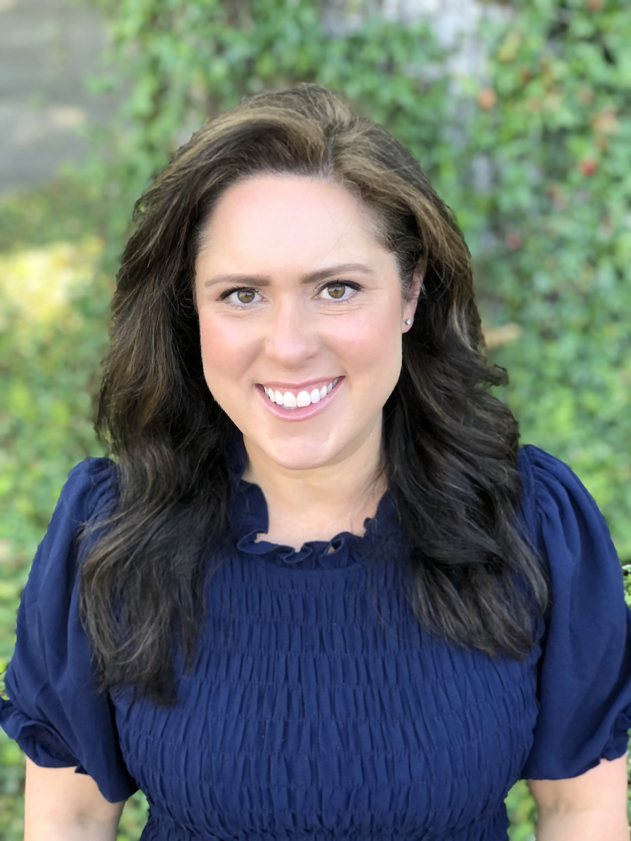 A woman with dark brown hair, light skin, and hazel eyes is smiling outdoors with a background of green foliage. She is wearing a blue blouse with a ruffled neckline.