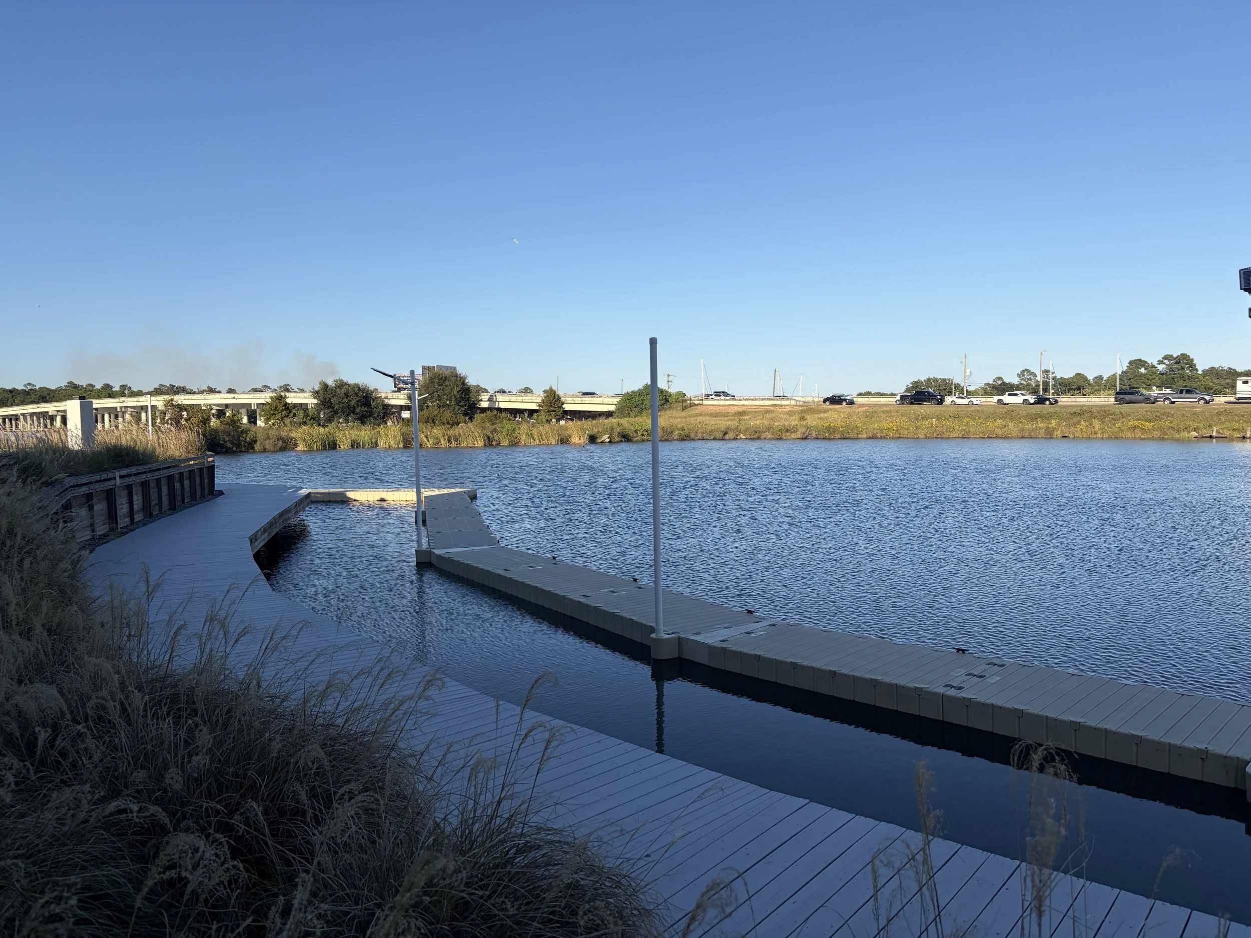 A waterfront scene with a wooden dock extending into a body of water, surrounded by grasses and a rail, under a clear blue sky. In the background, there are trees, a highway with moving cars, and sailboats docked along the shore.