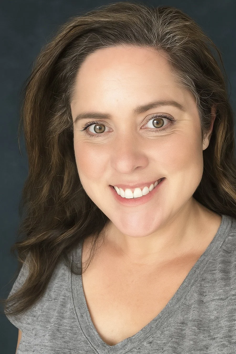Close-up portrait of a smiling woman with brown hair and hazel eyes, wearing a gray shirt.