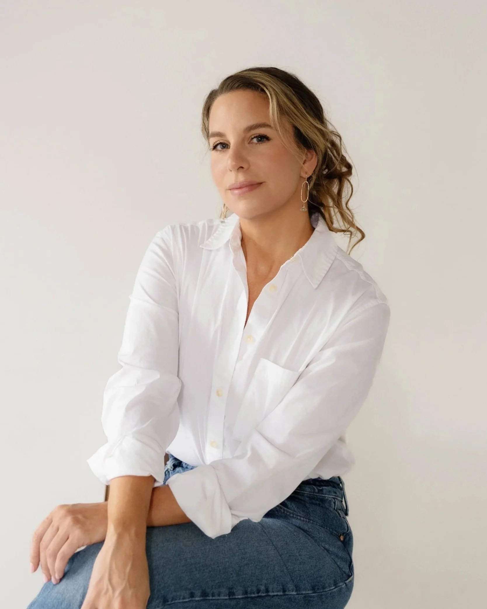 Woman with blonde hair wearing a white button-up shirt and blue jeans, sitting against a plain white wall.