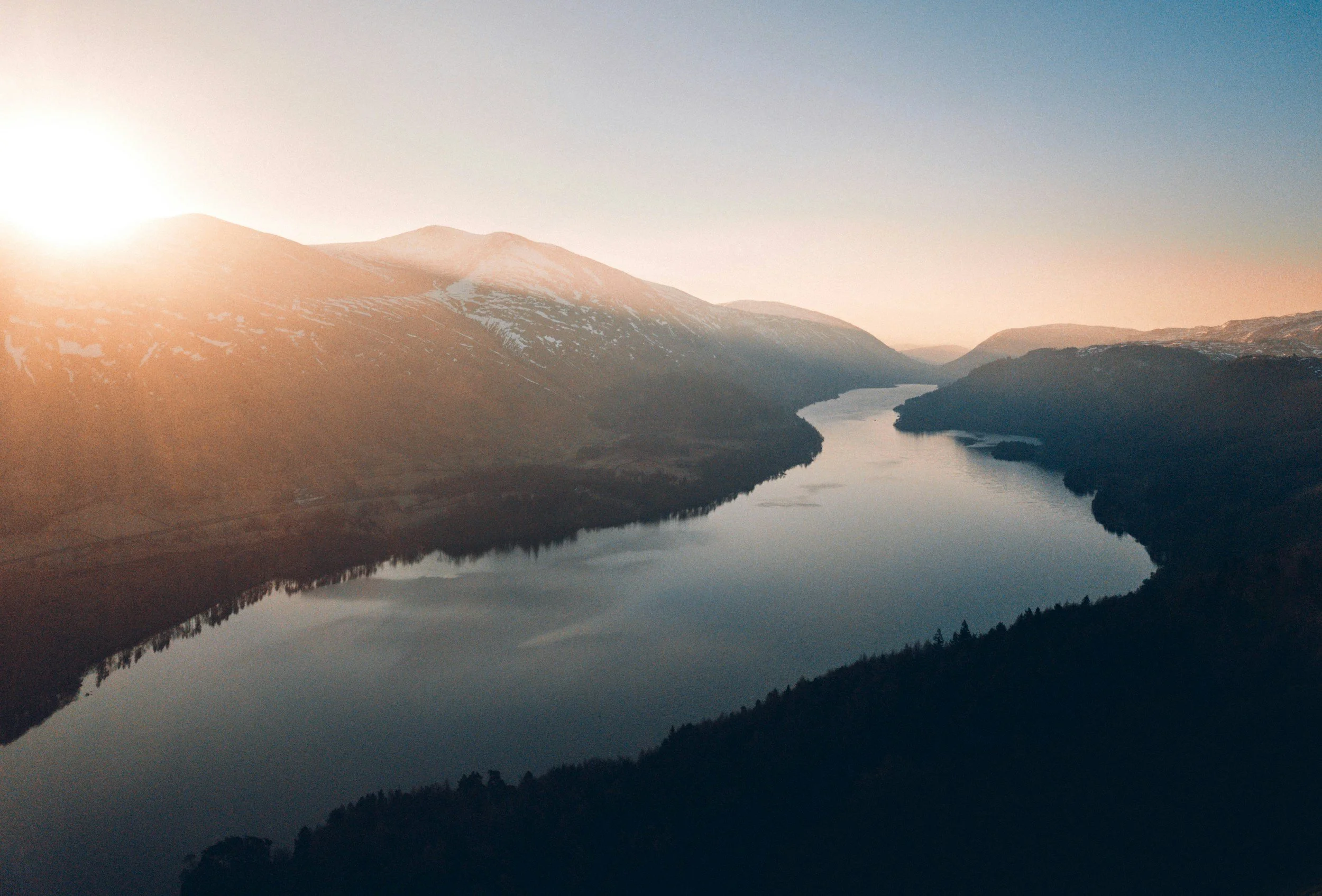 A wide view of a river winding through mountains at sunset, with snow patches on the peaks and a clear sky.