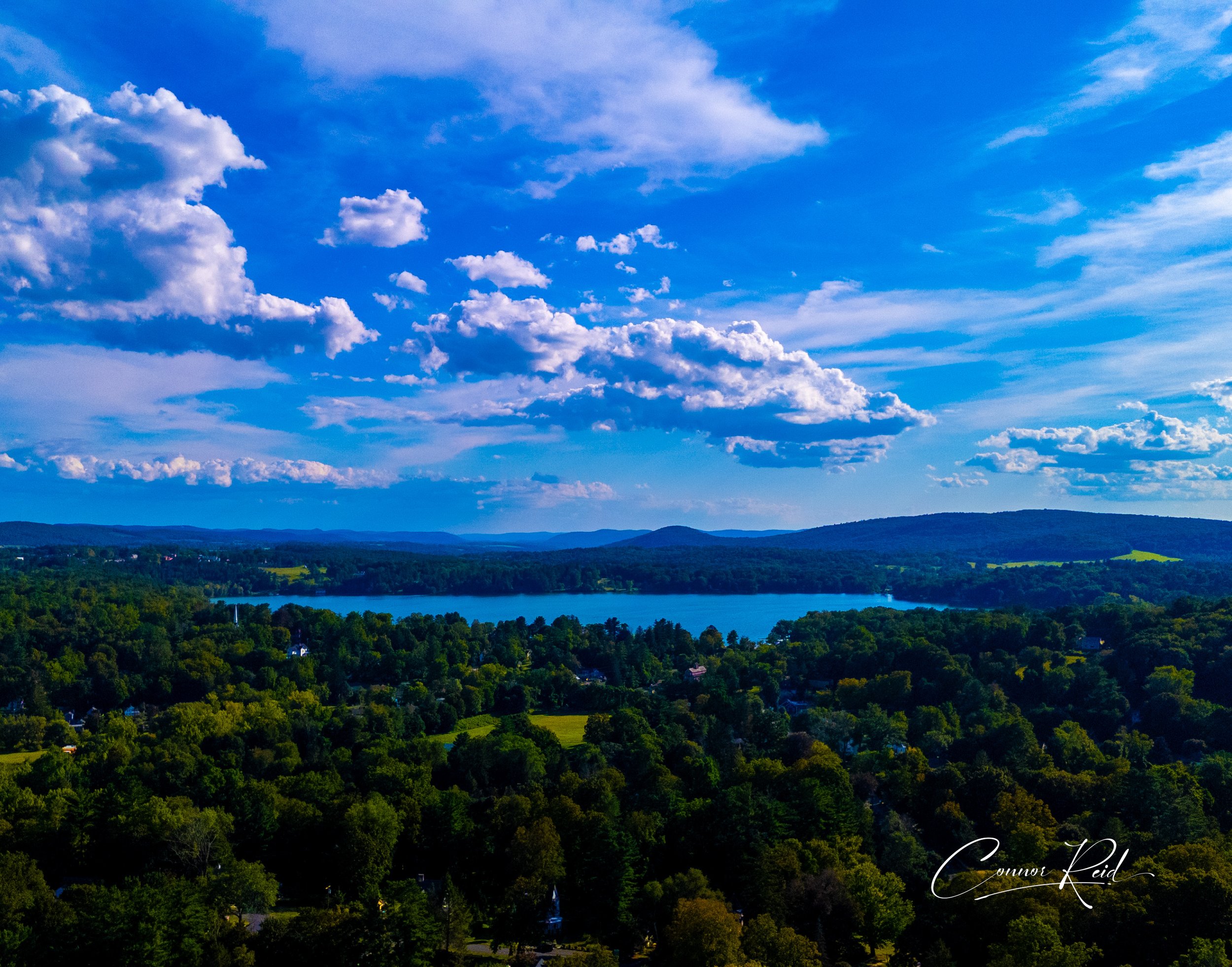 A scenic view of a large lake surrounded by lush green trees, with hills in the background under a cloudy blue sky.
