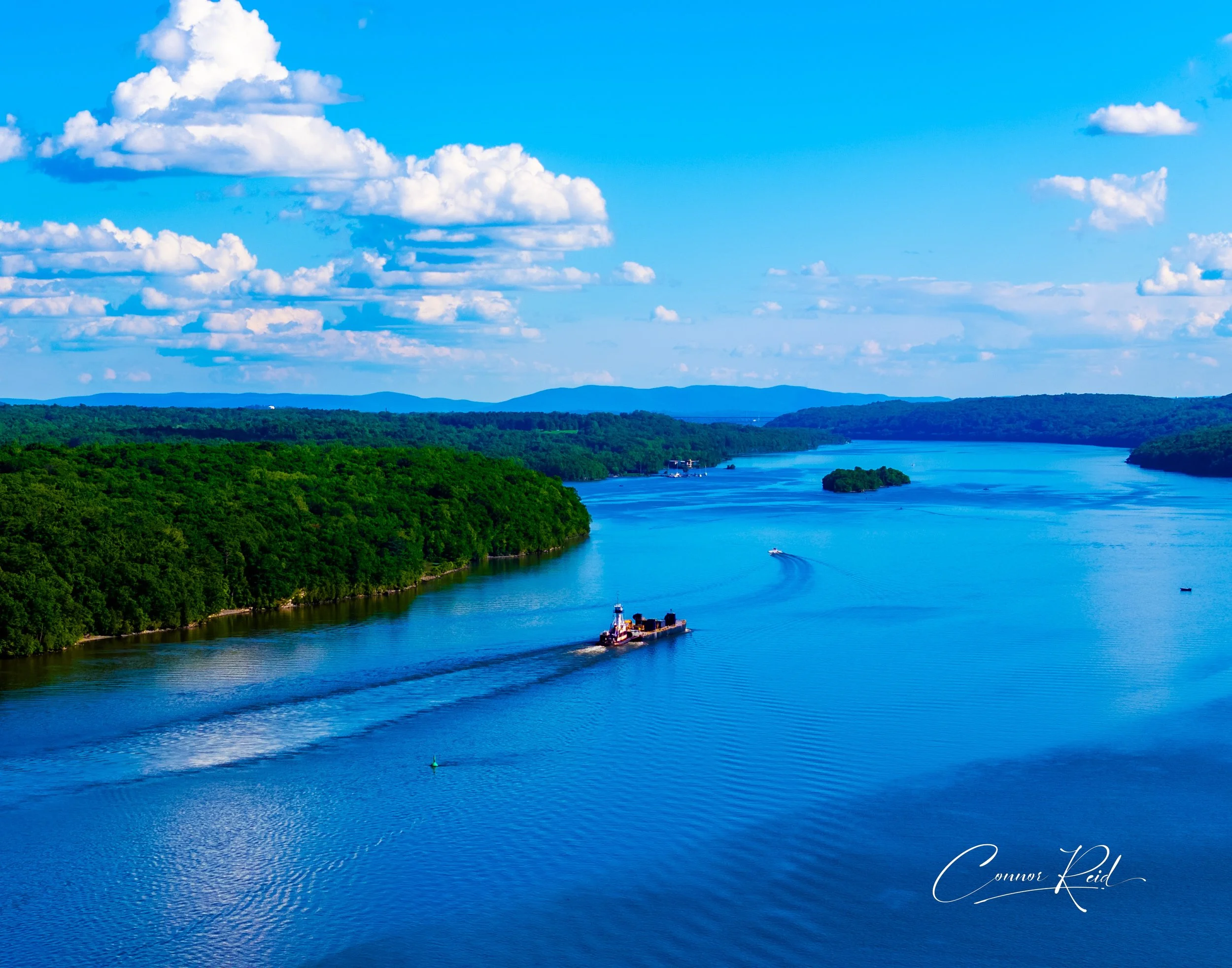 A scenic river landscape with lush green forests on both sides, blue water with three boats creating wakes, and a partly cloudy sky above.