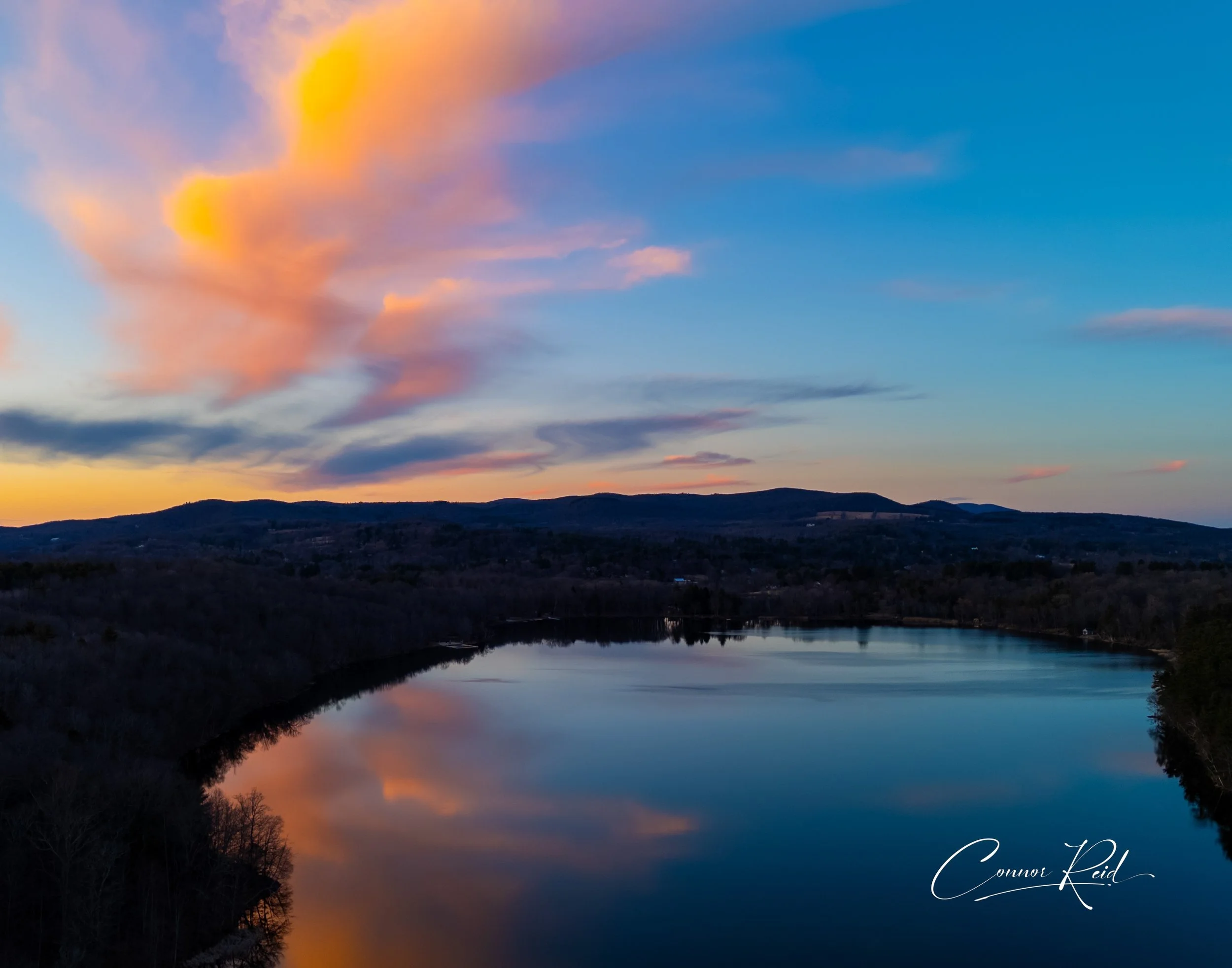 A scenic view of a river at sunset with colorful pink and orange clouds in the sky and mountain hills in the background.