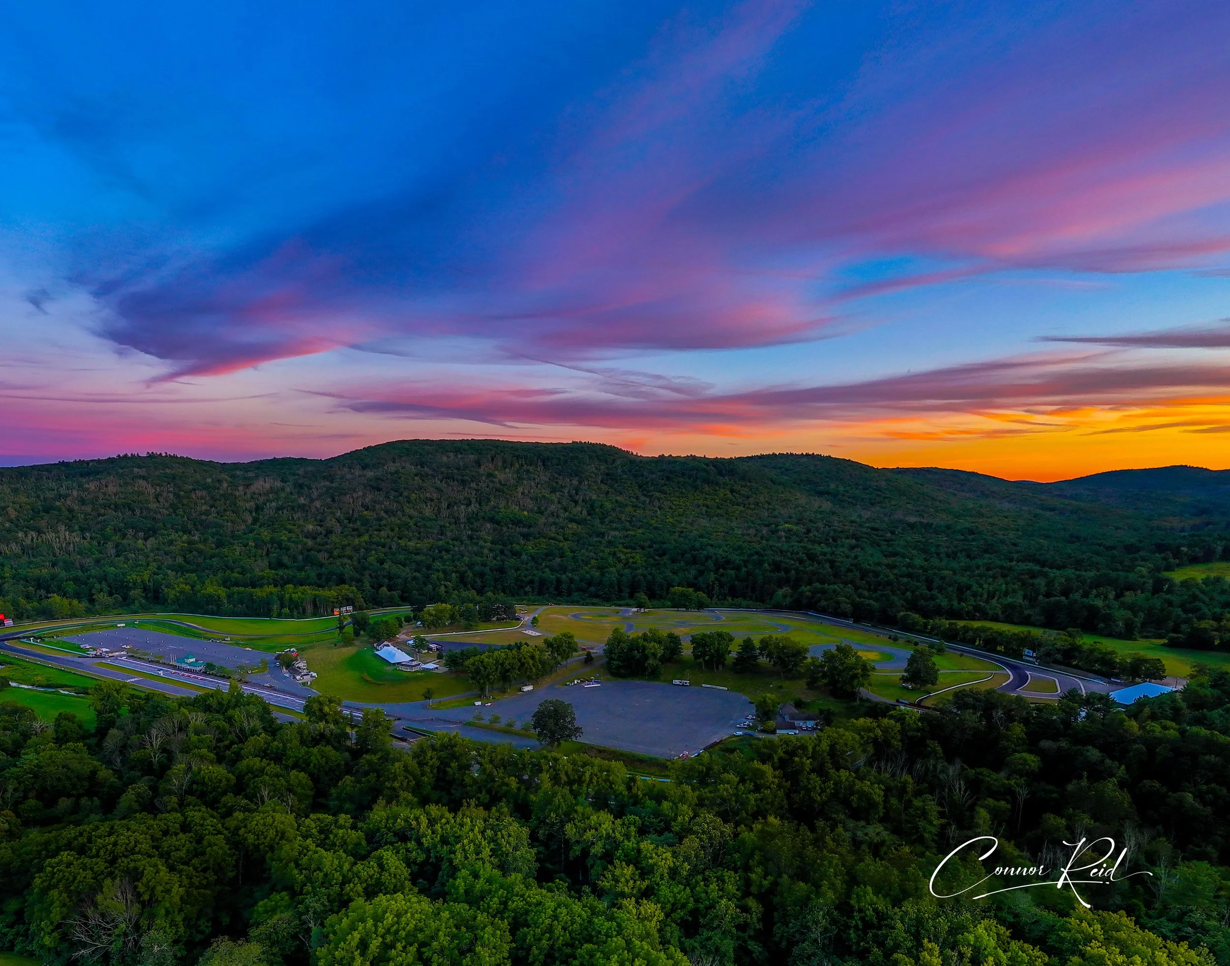 Colorful sunset over a lush green landscape with a racetrack and parking lot in the foreground.