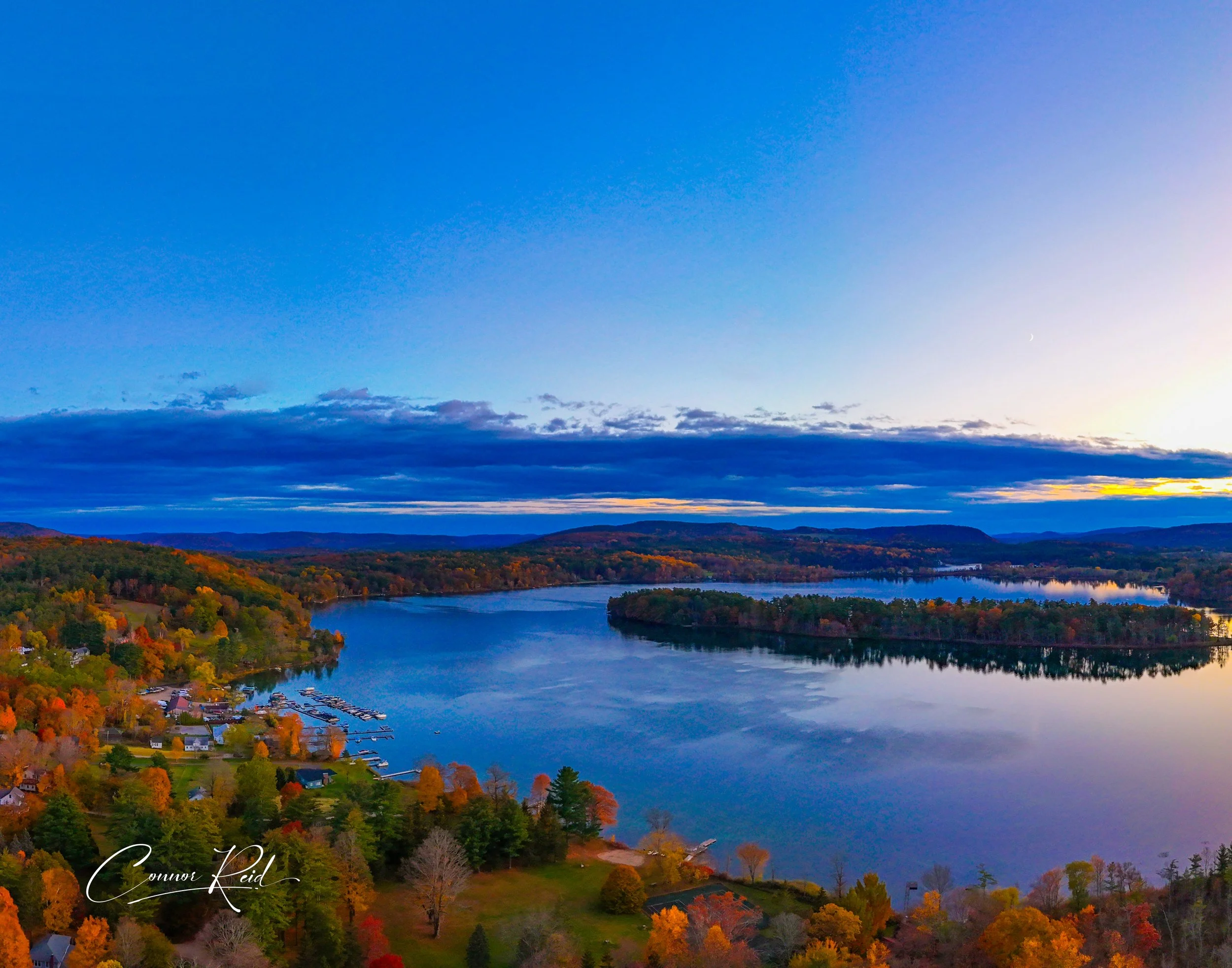 Aerial view of a lake surrounded by trees with fall foliage, a small marina, and a few houses under a partly cloudy sky at sunset.