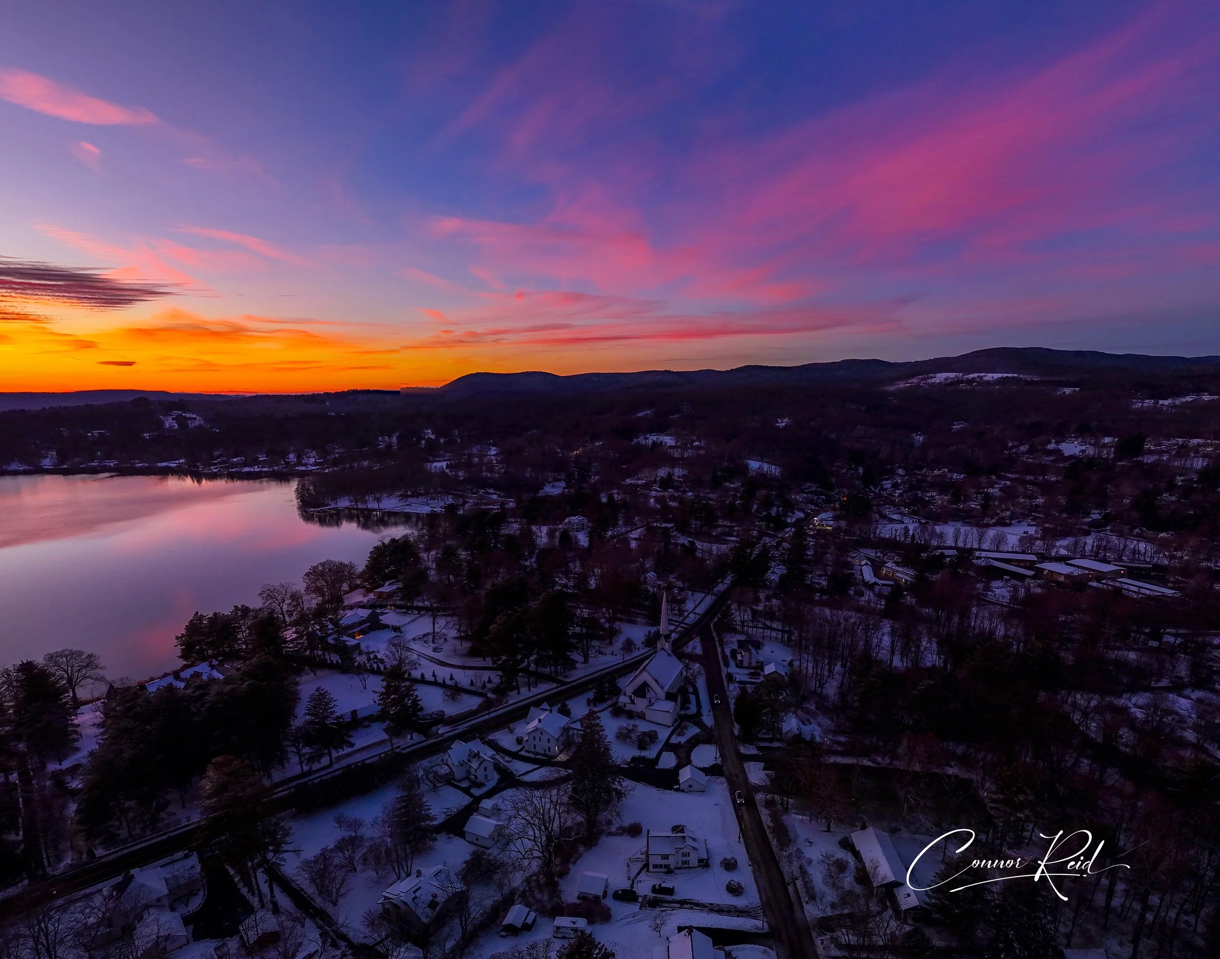 A winter sunset over a small town near a lake, with snow-covered houses and trees, colorful sky with pink and orange clouds, and distant mountains.