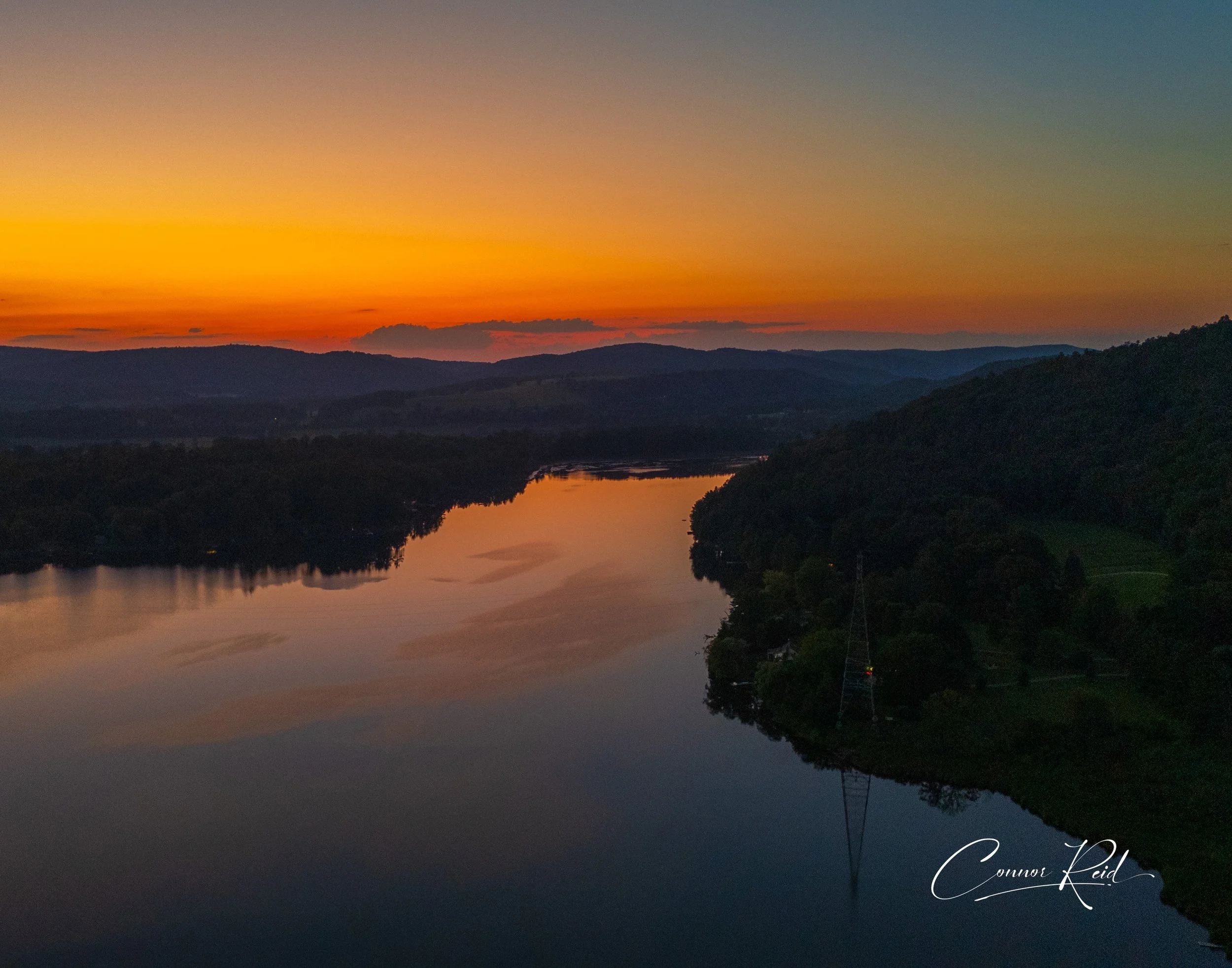 Sunset over a river with mountains in the background and tree-lined banks, with a reflection on the calm water.
