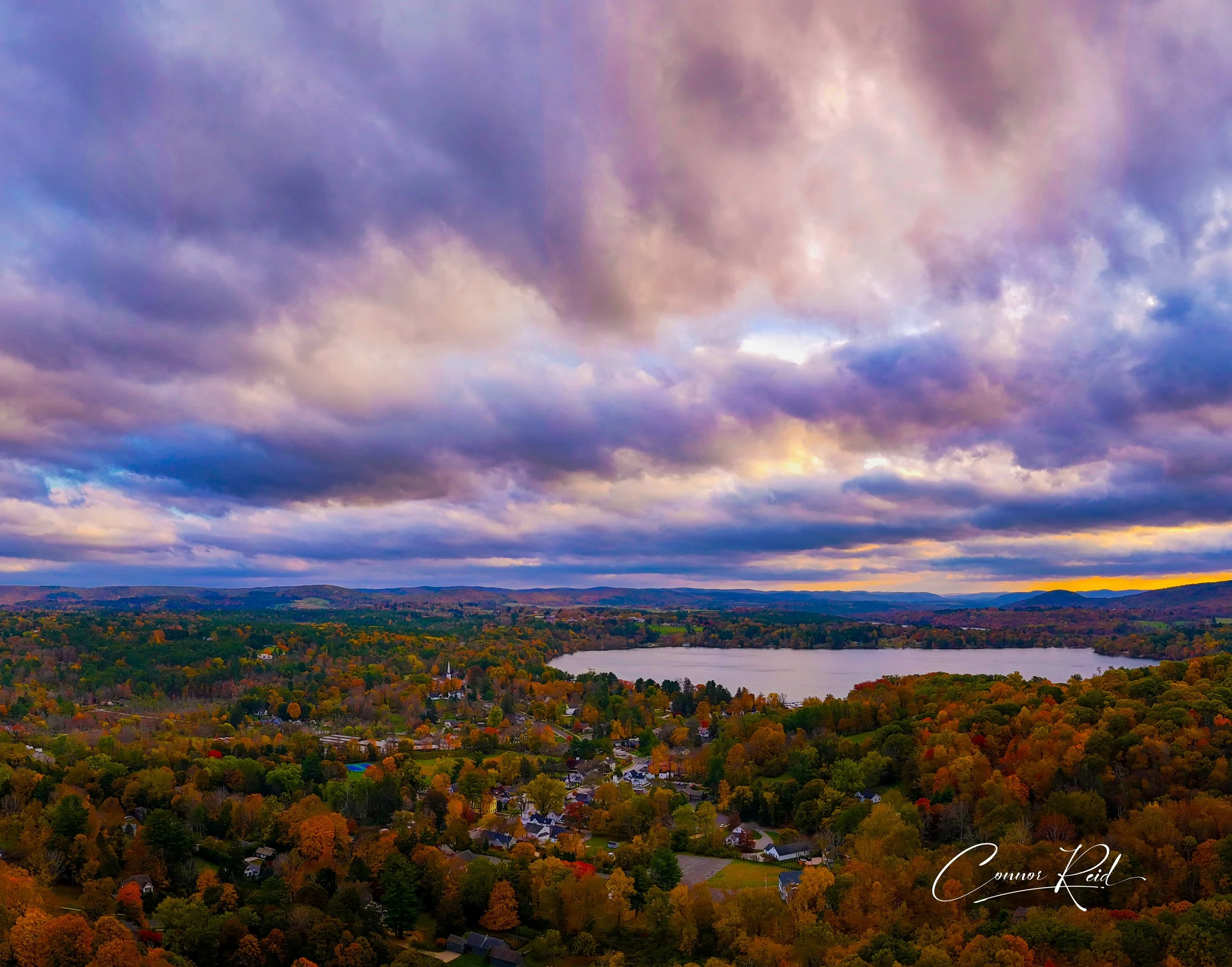 Aerial view of a lake surrounded by fall foliage trees with colorful leaves, a small town, and a cloudy sky at sunset.