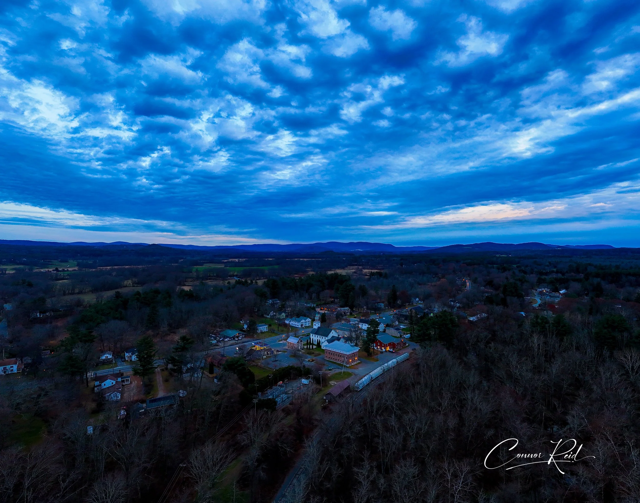 A landscape view of a small town surrounded by trees with rolling hills in the background under a cloudy blue sky during twilight.