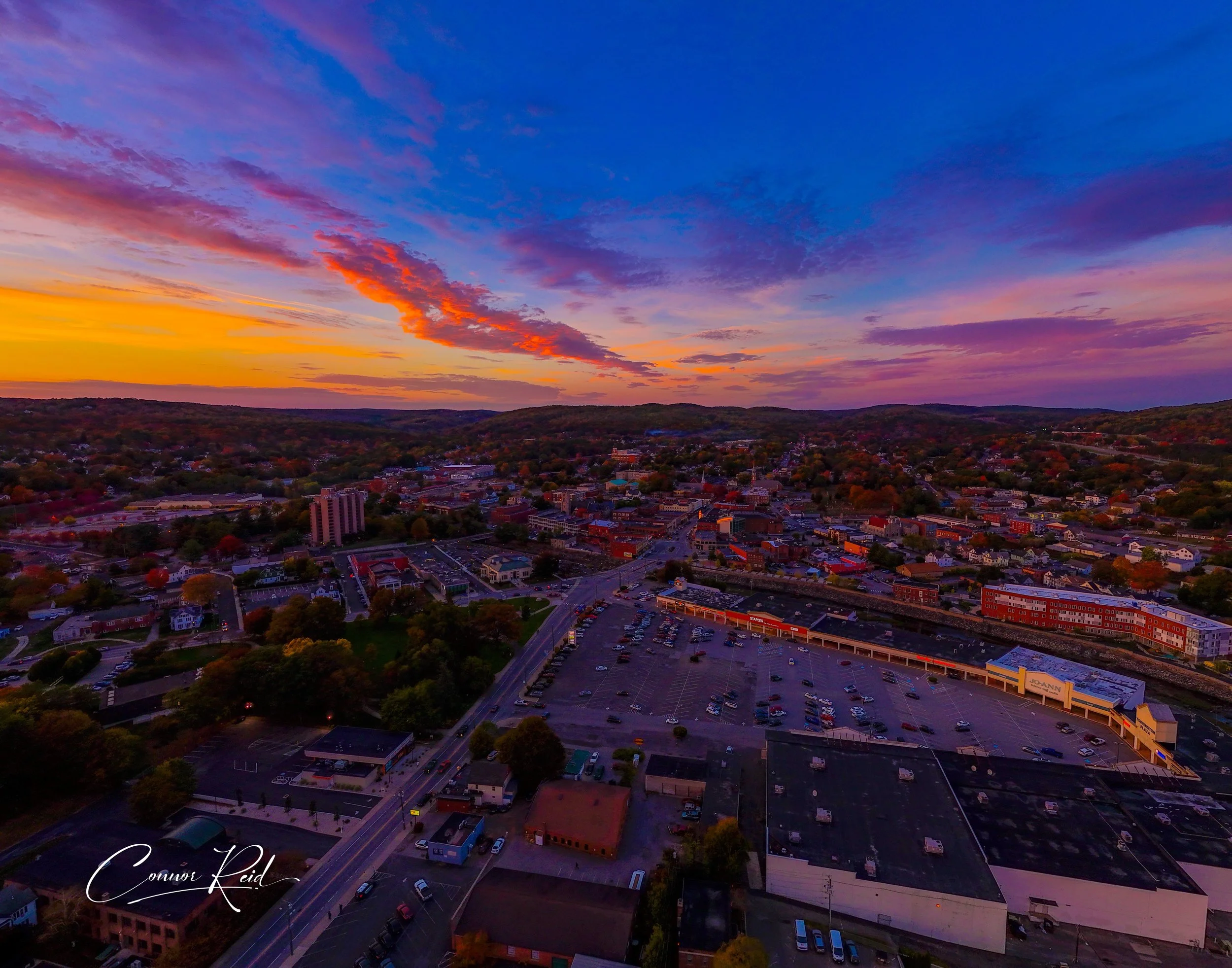 Aerial view of a small town at sunset with colorful sky, buildings, streets, and parking lots visible.