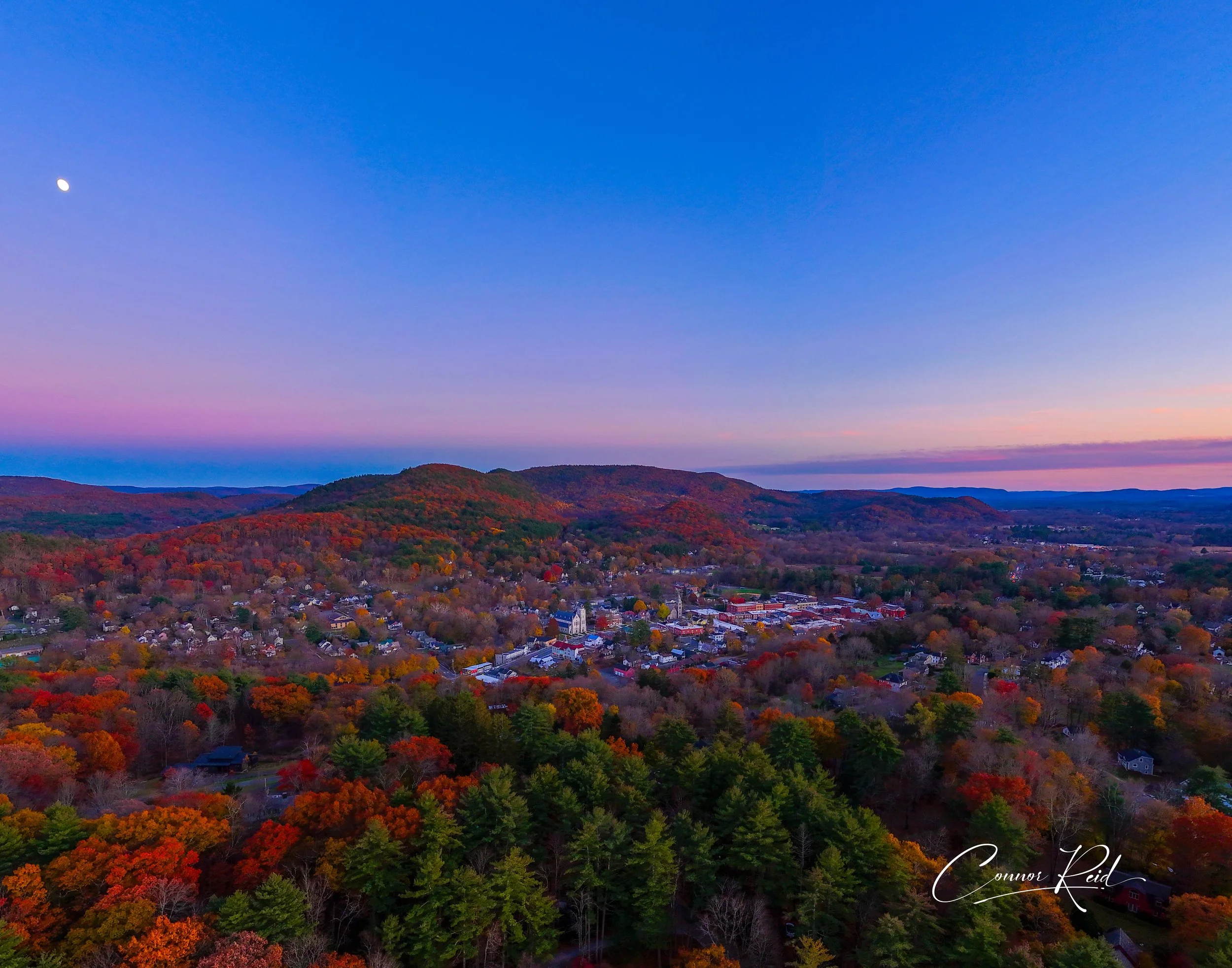 Aerial view of fall foliage in a small town with colorful trees, rolling hills, and a clear twilight sky with the moon overhead.