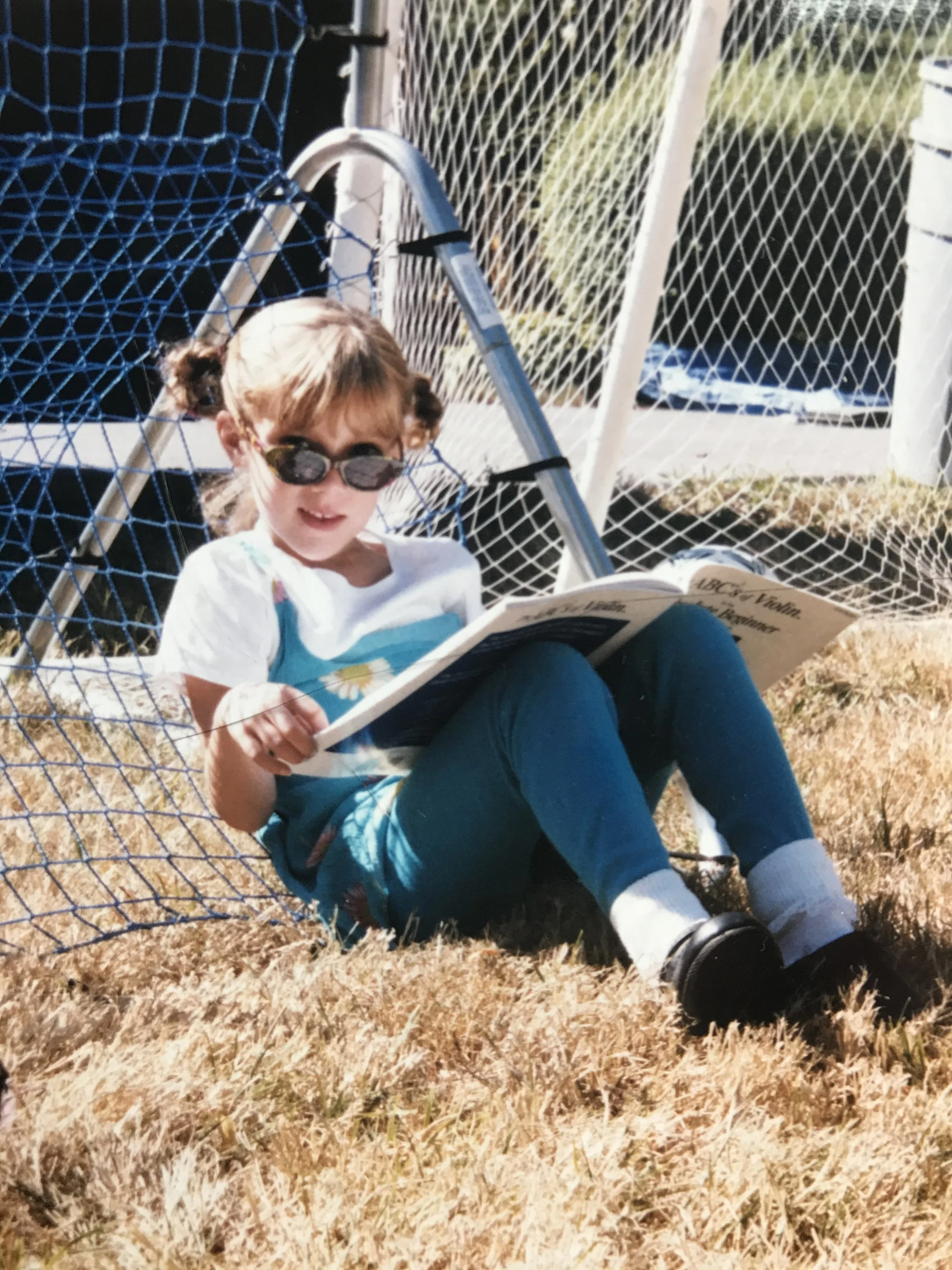 A young girl wearing sunglasses and a white shirt with a flower design is sitting on dry grass, reading a book.