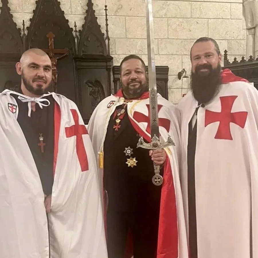 Three men dressed as knights with medieval-style capes and crosses, standing in a church, one holding a sword, smiling at the camera.