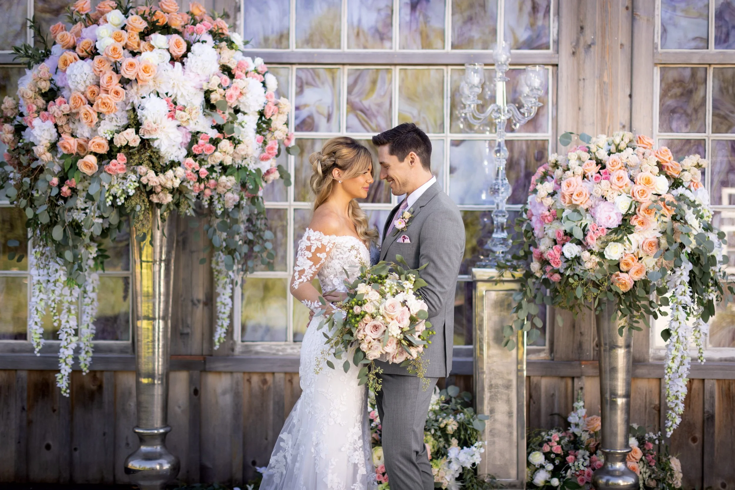 A bride and groom stand close together at their wedding ceremony, gazing into each other's eyes, surrounded by large pink and white floral arrangements and tall vases, with a rustic wooden backdrop and window panes.