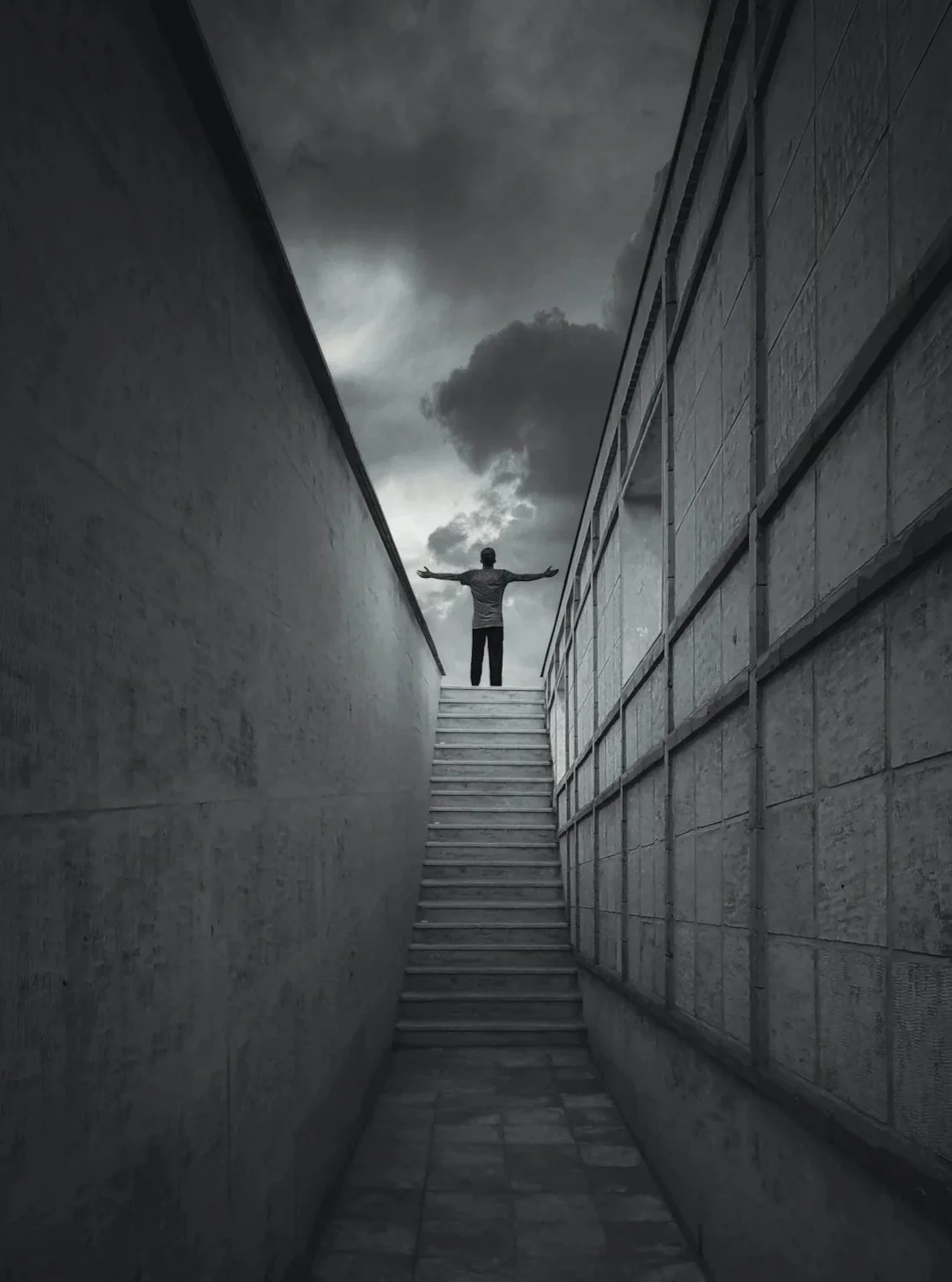 A person with arms outstretched standing at the top of a staircase between two dark, narrow walls, looking at a cloudy sky.