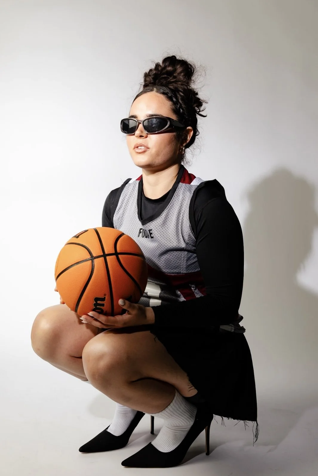 A woman in black high heels, white socks, and black clothing crouches while holding a basketball, wearing dark sunglasses, a gray sports jersey, and her hair in a messy bun against a plain white background.
