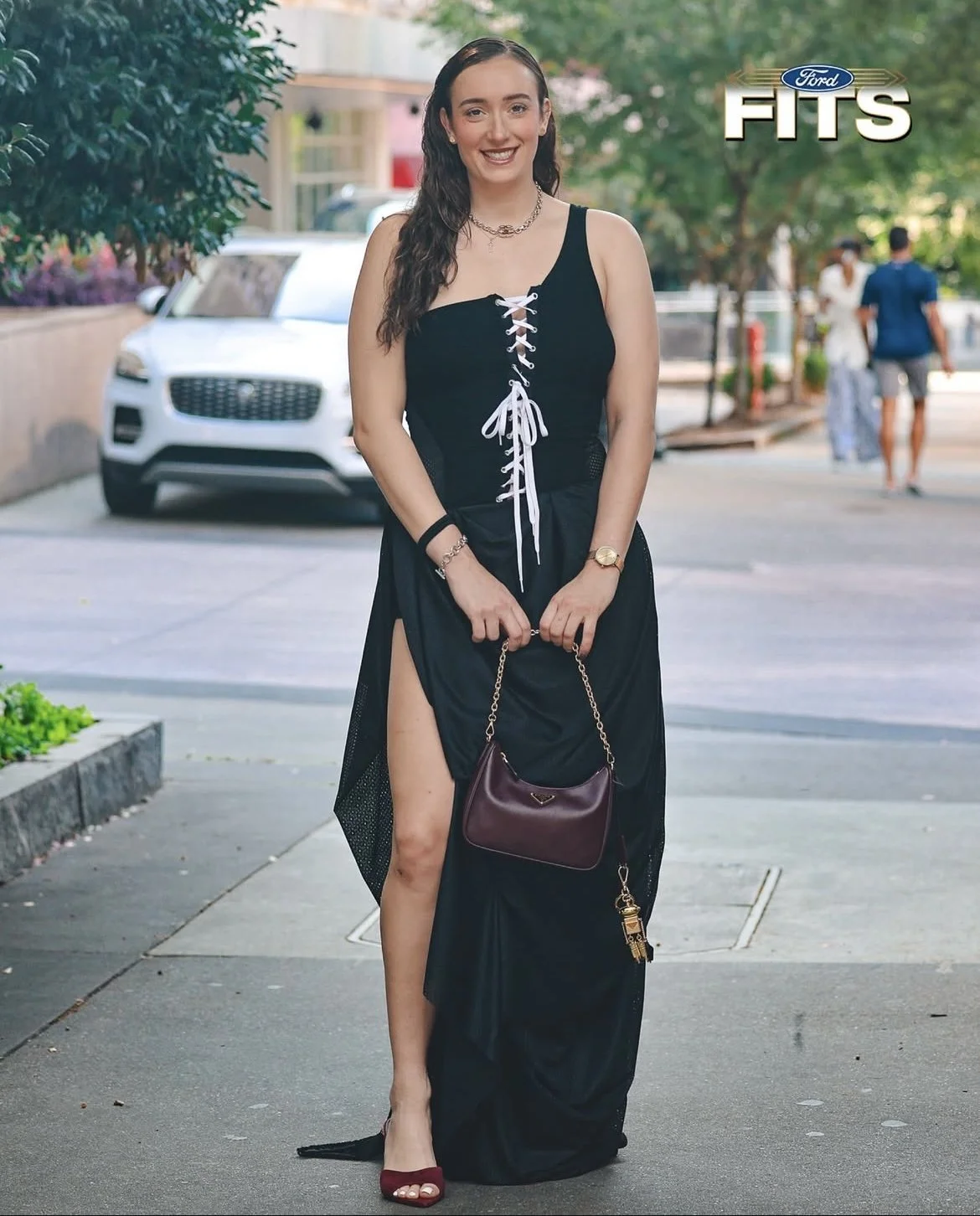 A young woman with wavy brown hair, smiling, wearing a long black dress with slits, red high heels, holding a small burgundy handbag with a gold chain, standing on a city sidewalk with trees and cars in the background.