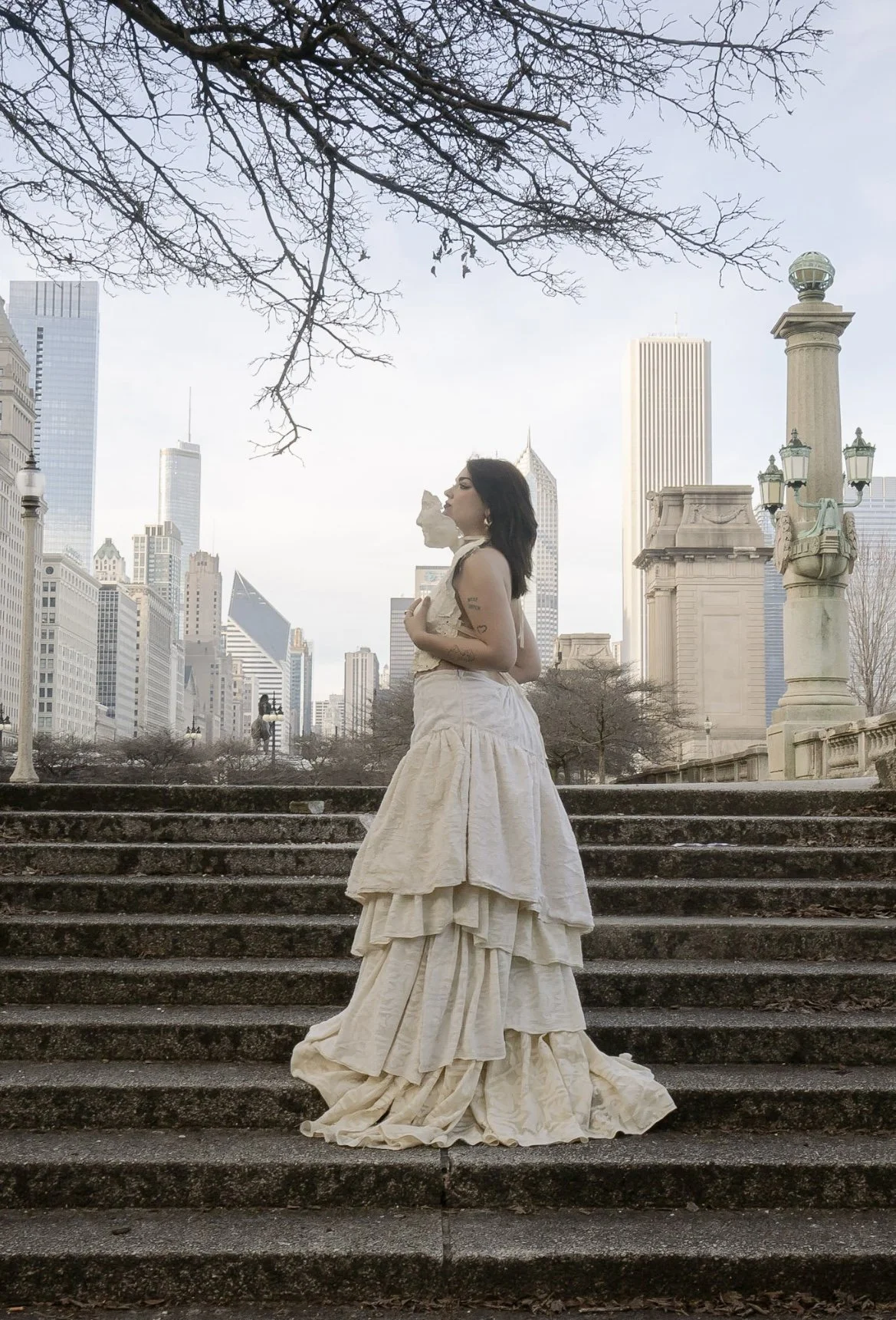 A woman in a vintage cream-colored dress stands on stone stairs in a city park, with tall skyscrapers in the background and leafless tree branches overhead.
