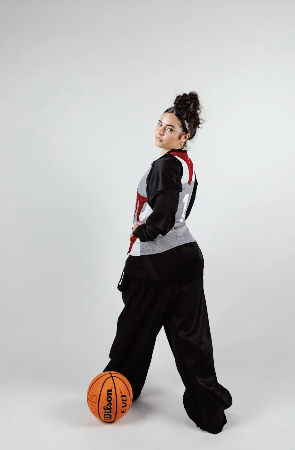 A woman standing beside a basketball, wearing a black, red, and white sports jersey and black loose-fitting pants, with her hair styled in a high bun, against a plain light background.
