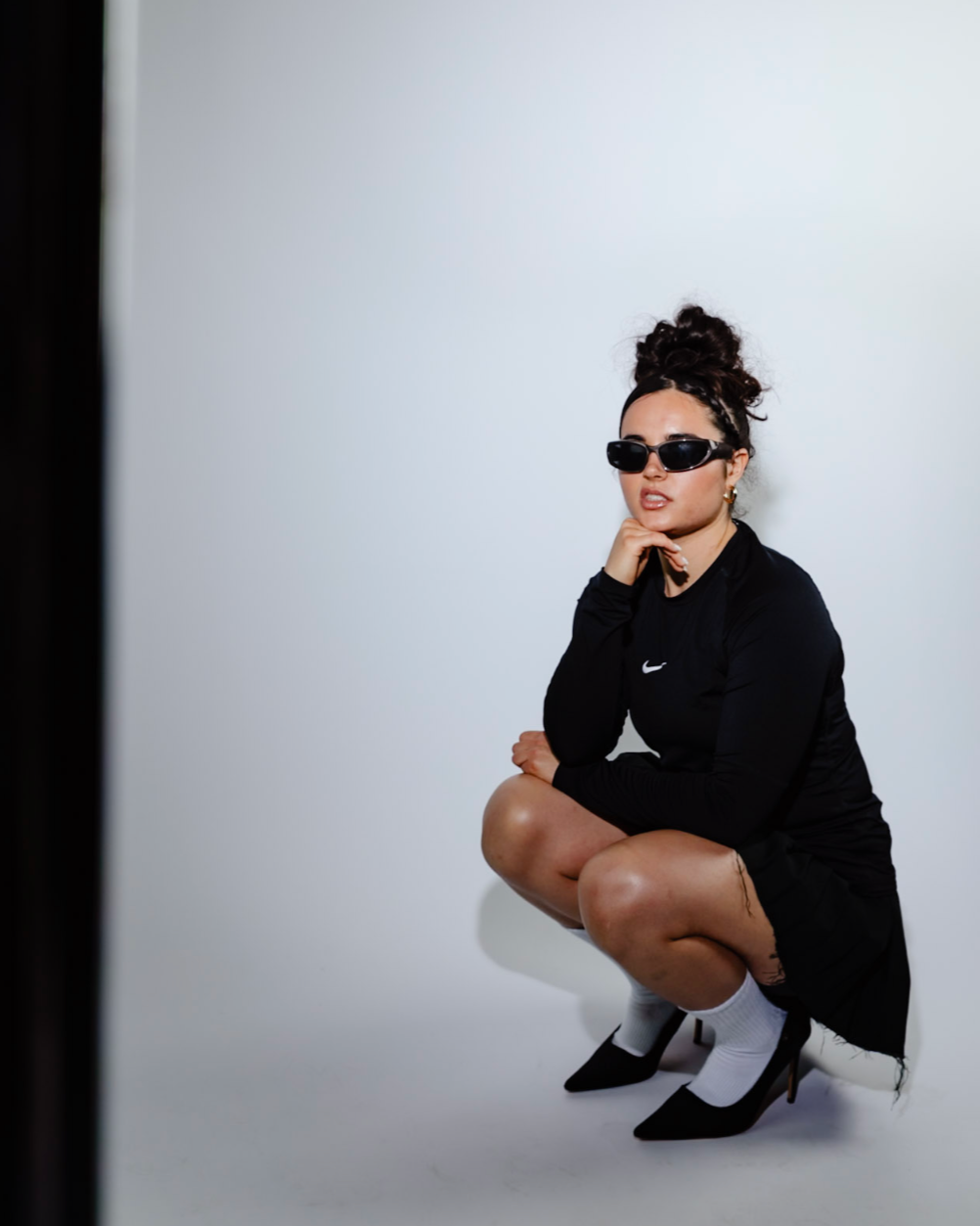 A woman squatting against a white background, wearing black Nike athletic wear, black high heels, white socks, sunglasses, hoop earrings, and a bag on her shoulder.
