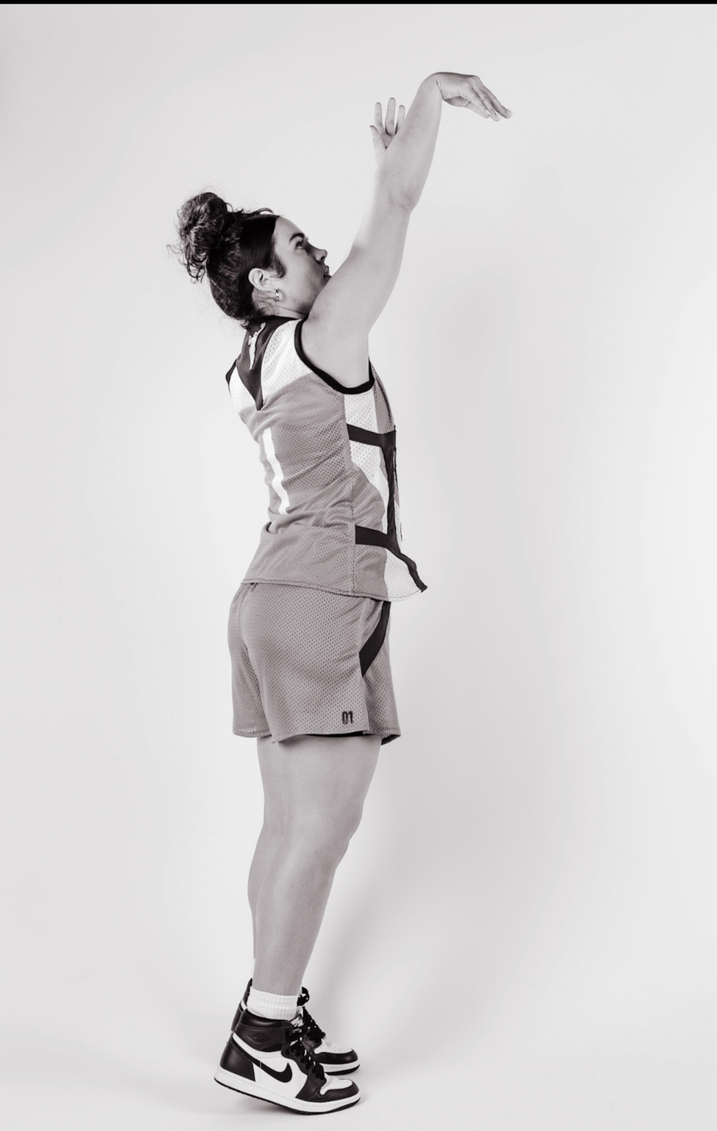 A woman in athletic shorts, T-shirt, and sneakers stretching with arms raised above her head against a plain background.