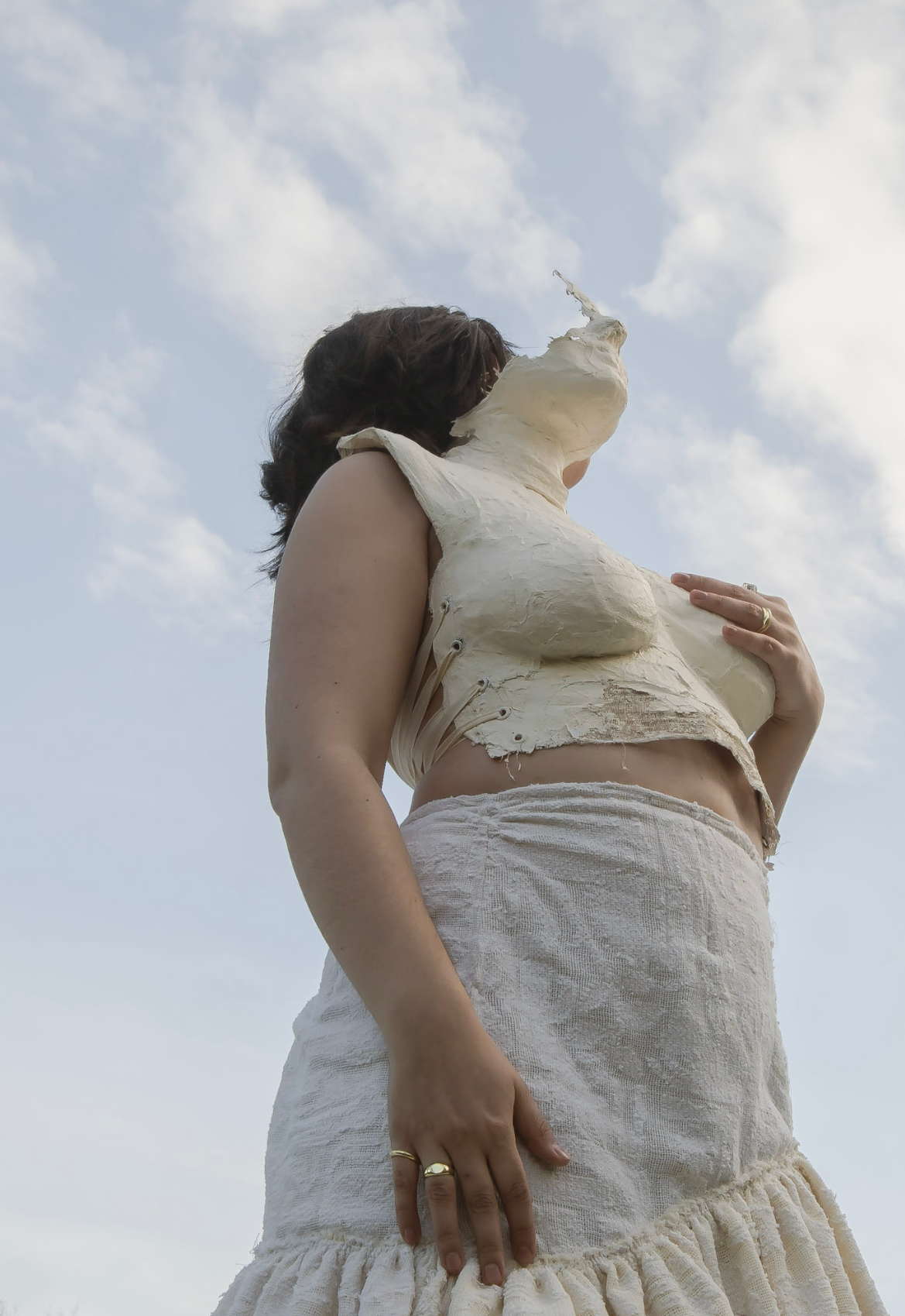 A person with dark hair wearing a cream-colored, textured skirt and a cropped top made of wax-like material, standing outdoors against a cloudy sky.