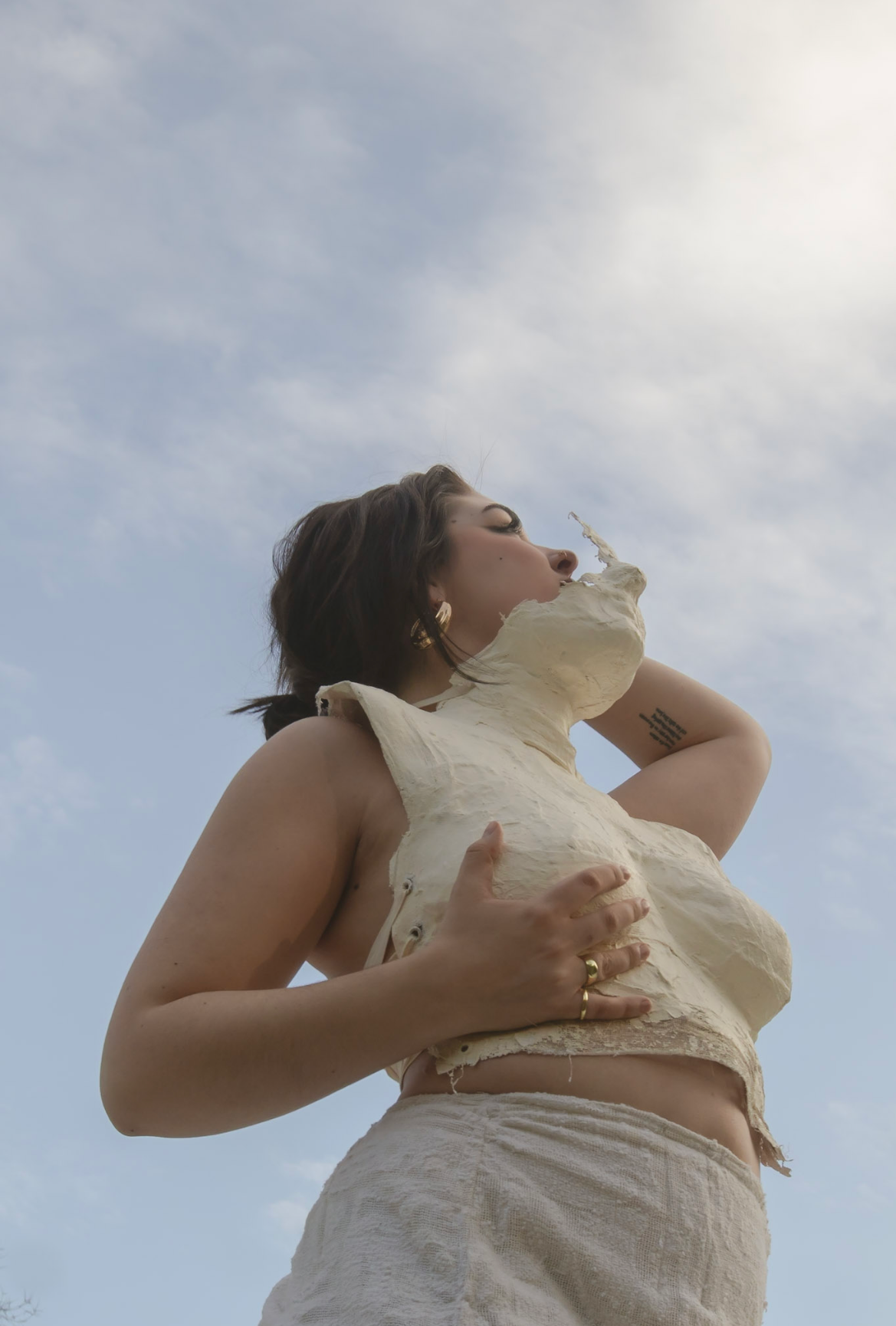 A woman standing outdoors under a cloudy sky, wearing a cream-colored, torn, sleeveless top and matching shorts, holding her large ceramic sculpture with both hands.
