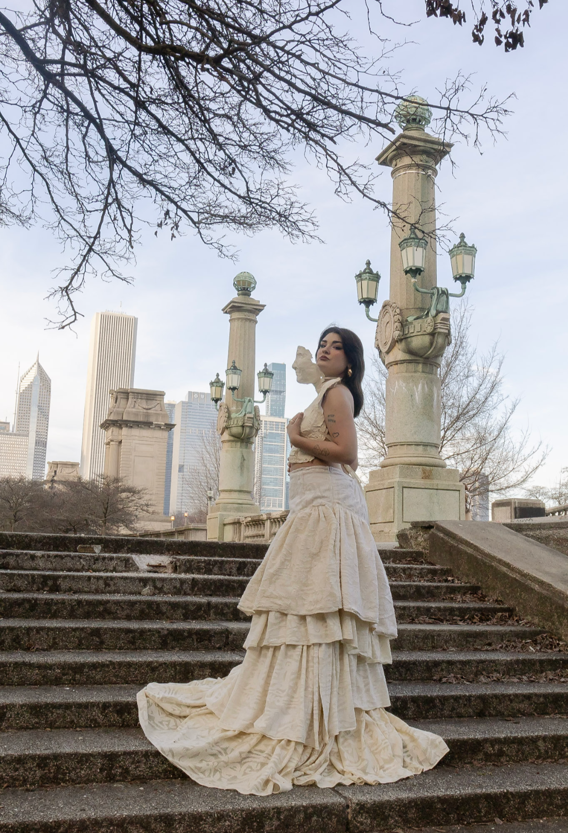 A woman with dark hair and tattoos wearing a beige, layered, vintage dress stands on stone steps in an outdoor park with city skyscrapers in the background. She is looking over her shoulder with a confident expression, under leafless tree branches an