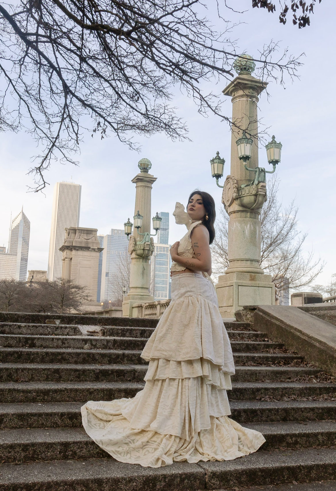A woman in a vintage cream-colored dress with ruffles stands on stone steps in a park with a city skyline in the background. She has dark hair, makeup, and tattoos on her arm, with leafless trees surrounding her.