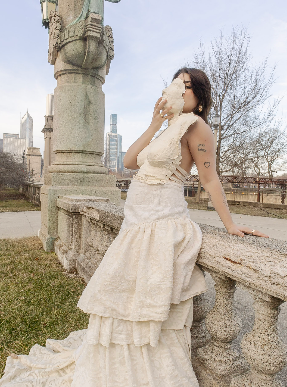 A woman wearing a light-colored dress sitting on a stone ledge in an outdoor urban setting during daytime, holding a large piece of bread or pastry near her face, with city skyscrapers and leafless trees in the background.