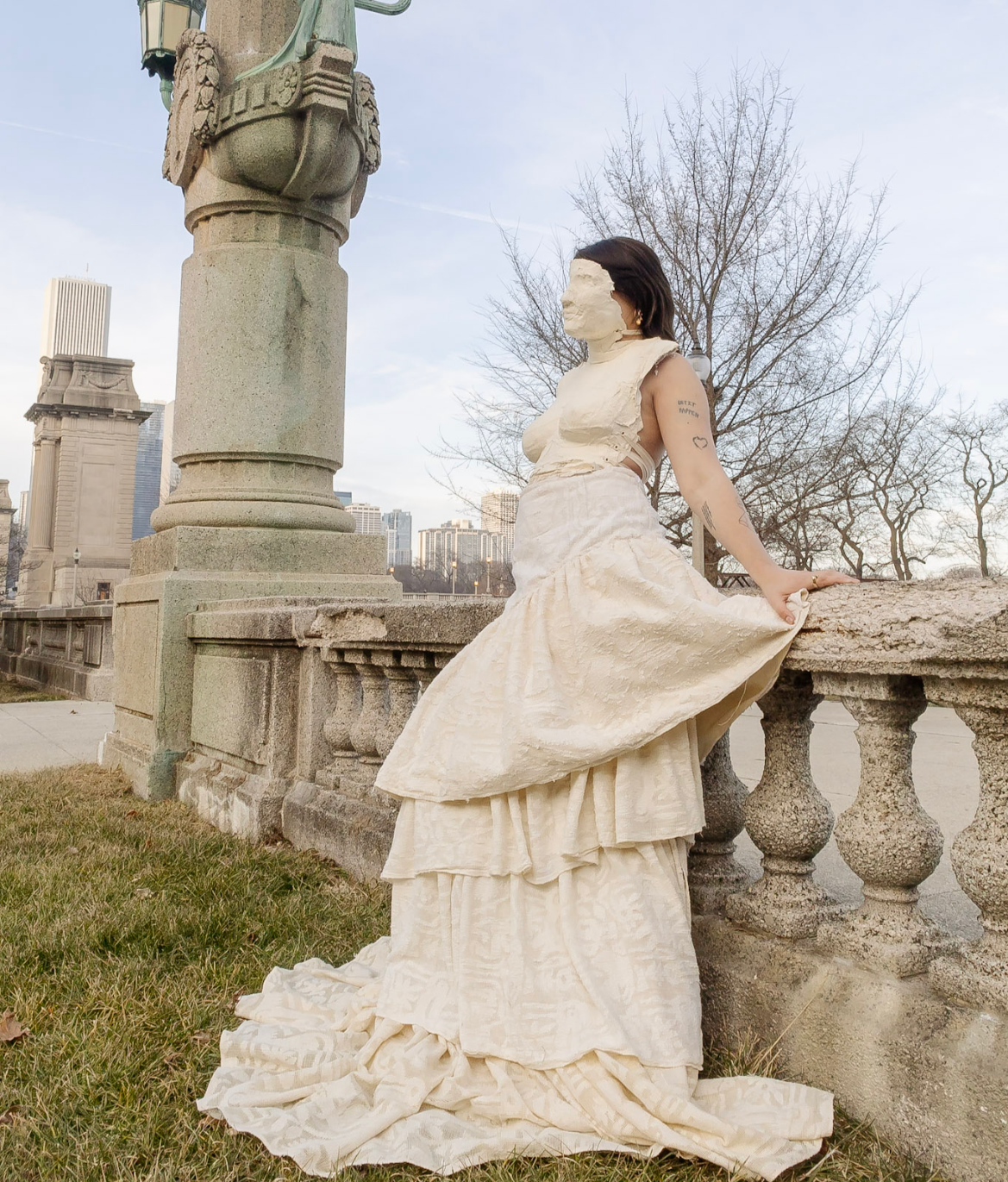 A woman in a cream-colored, layered, vintage-style dress with a long train, leaning back on a stone balustrade in an outdoor park or cityscape with tall buildings and leafless trees in the background.