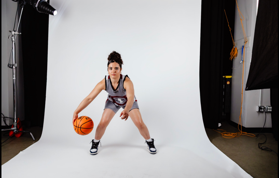 A young woman in a basketball jersey and shorts is posing with a basketball in a photography studio with a white backdrop, surrounded by studio lights and equipment.