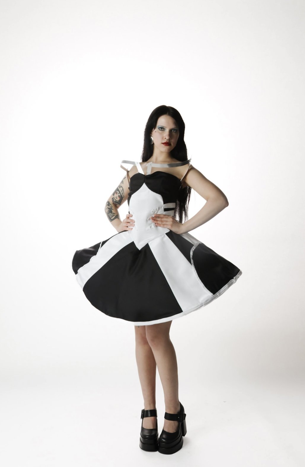 A woman with long dark hair, tattoos on her left arm, and makeup wearing a black and white dress with a full skirt, standing with her hands on her hips in front of a plain white background.